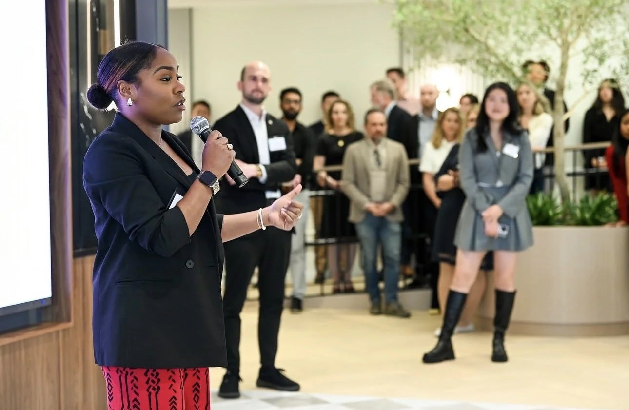 Jessicah Pierre in a black blazer and red pants speaks into a microphone at a professional event, with an audience of diverse men and women listening attentively.