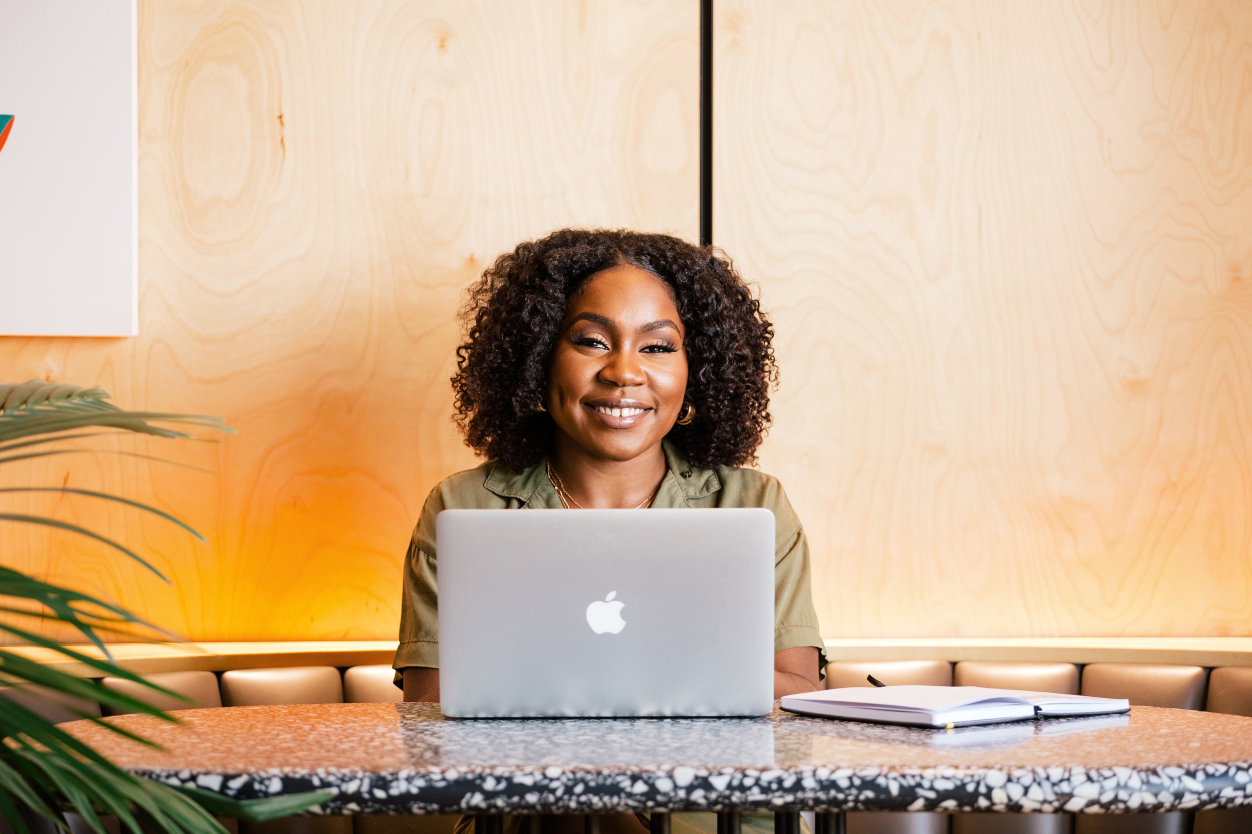 A woman with curly hair sitting at a table with a silver MacBook laptop in front of her, smiling, with a notebook and pen on the right, in a modern office setting.