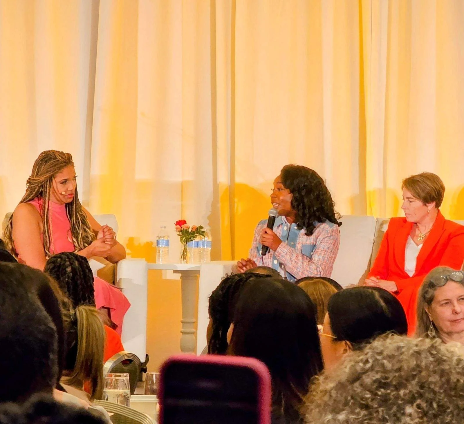 Three women seated on stage during a panel discussion, with an audience in front. The woman in the middle is speaking into a microphone. There are water bottles and small flowers on a table behind them, with yellow curtains in the background.