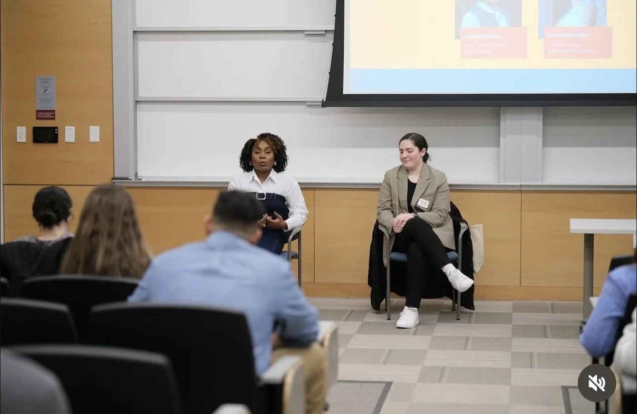 Two women seated at the front of a classroom or lecture hall, speaking to an audience of students.