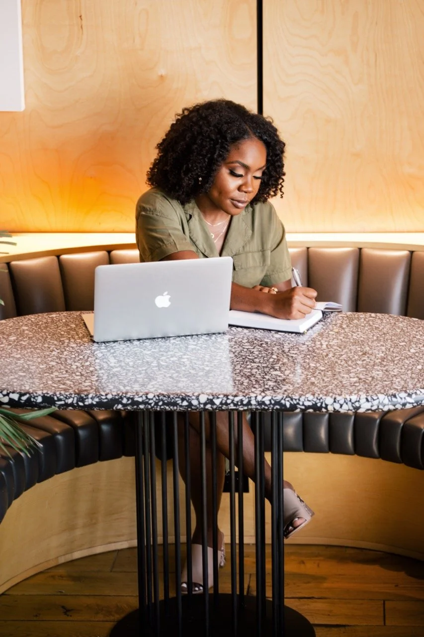 Jessicah Pierre with curly hair and olive green shirt sitting at a marble-topped table with a laptop and notebook, in a modern, warmly lit cafe or restaurant setting.