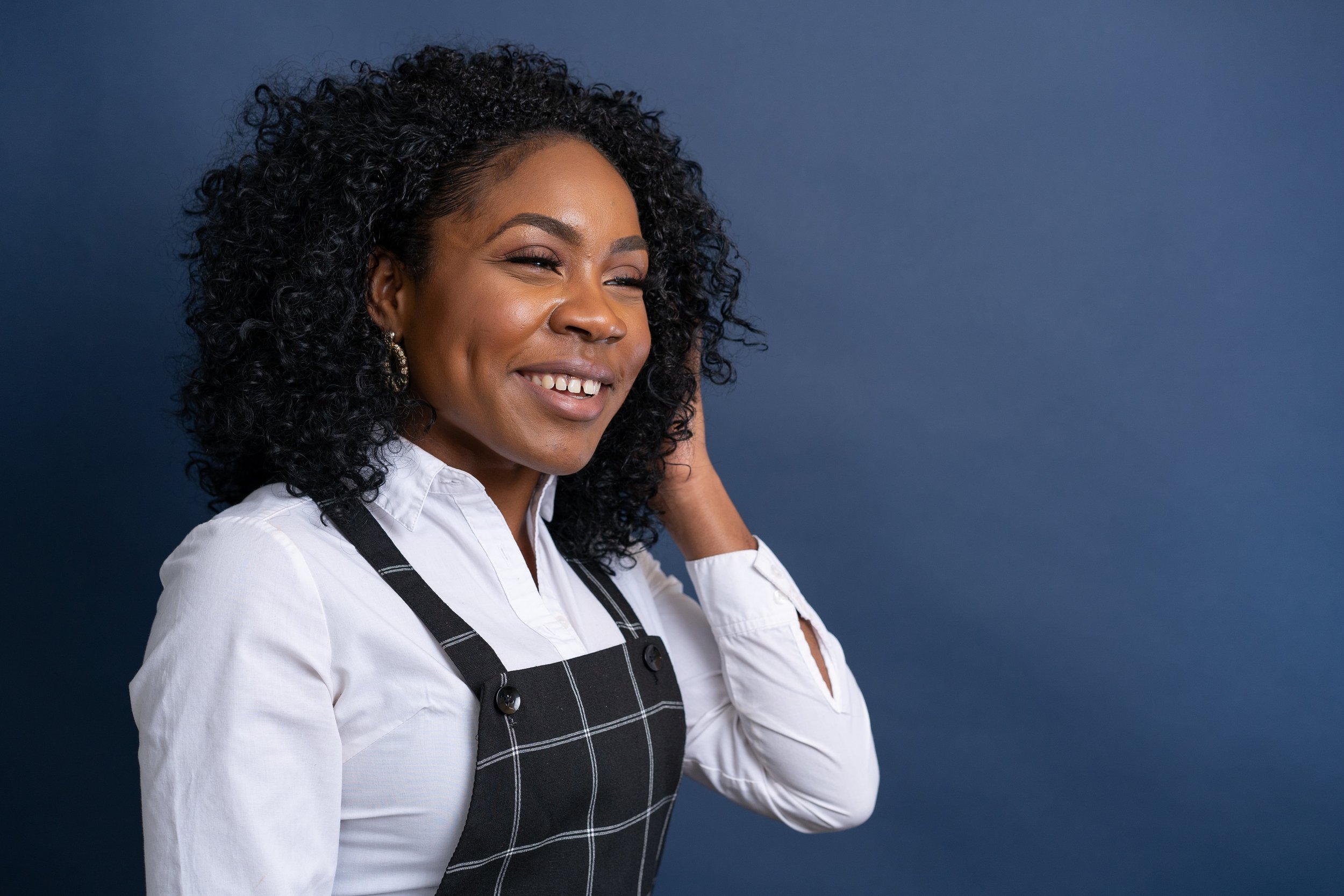 A woman with curly black hair smiling, wearing a white shirt and black apron with a white grid pattern, against a dark blue background.