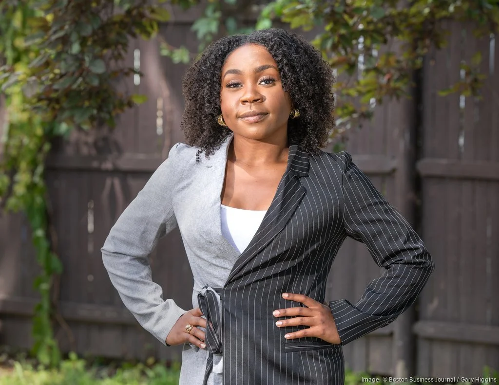 A woman stands outdoors in front of a wooden fence with greenery, wearing a suit with one half being light gray and the other half black with pinstripes, posing confidently with her hands on her hips.