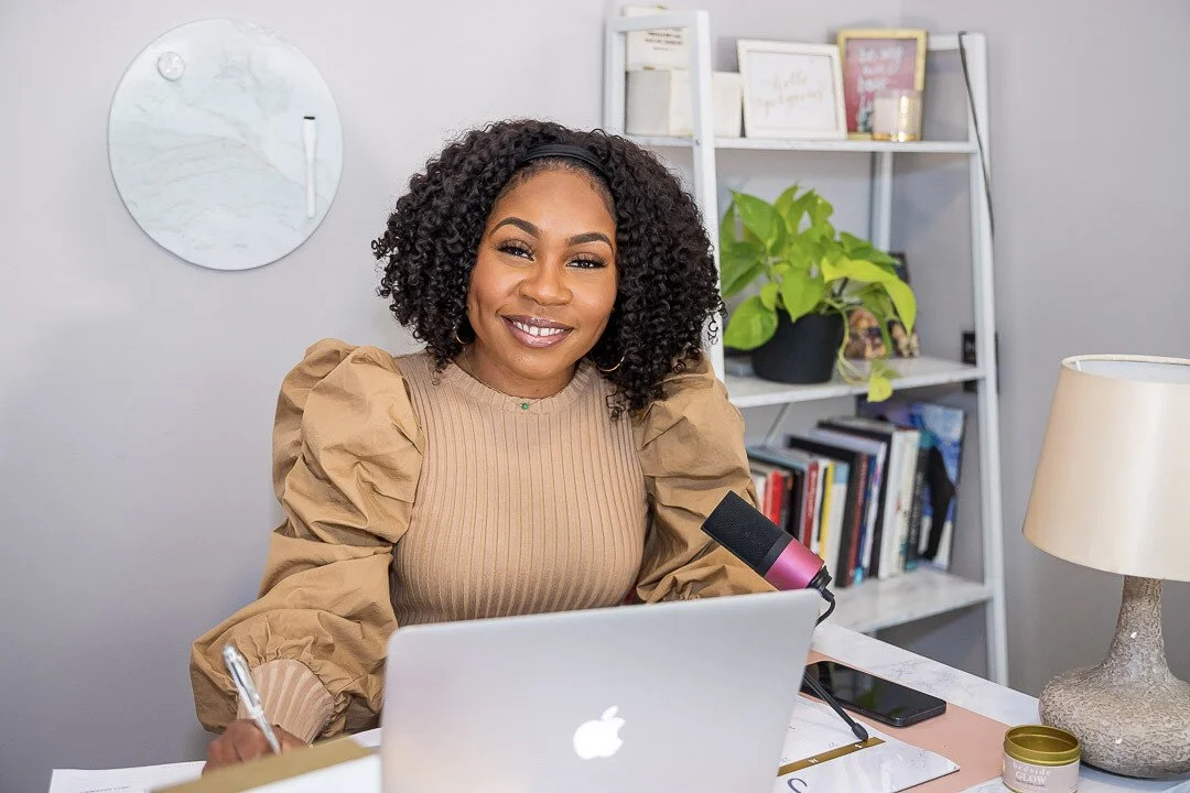 Jessicah Pierre sitting at a desk with a laptop, smiling at the camera, in a room with a bookshelf, potted plant, and decorative items.