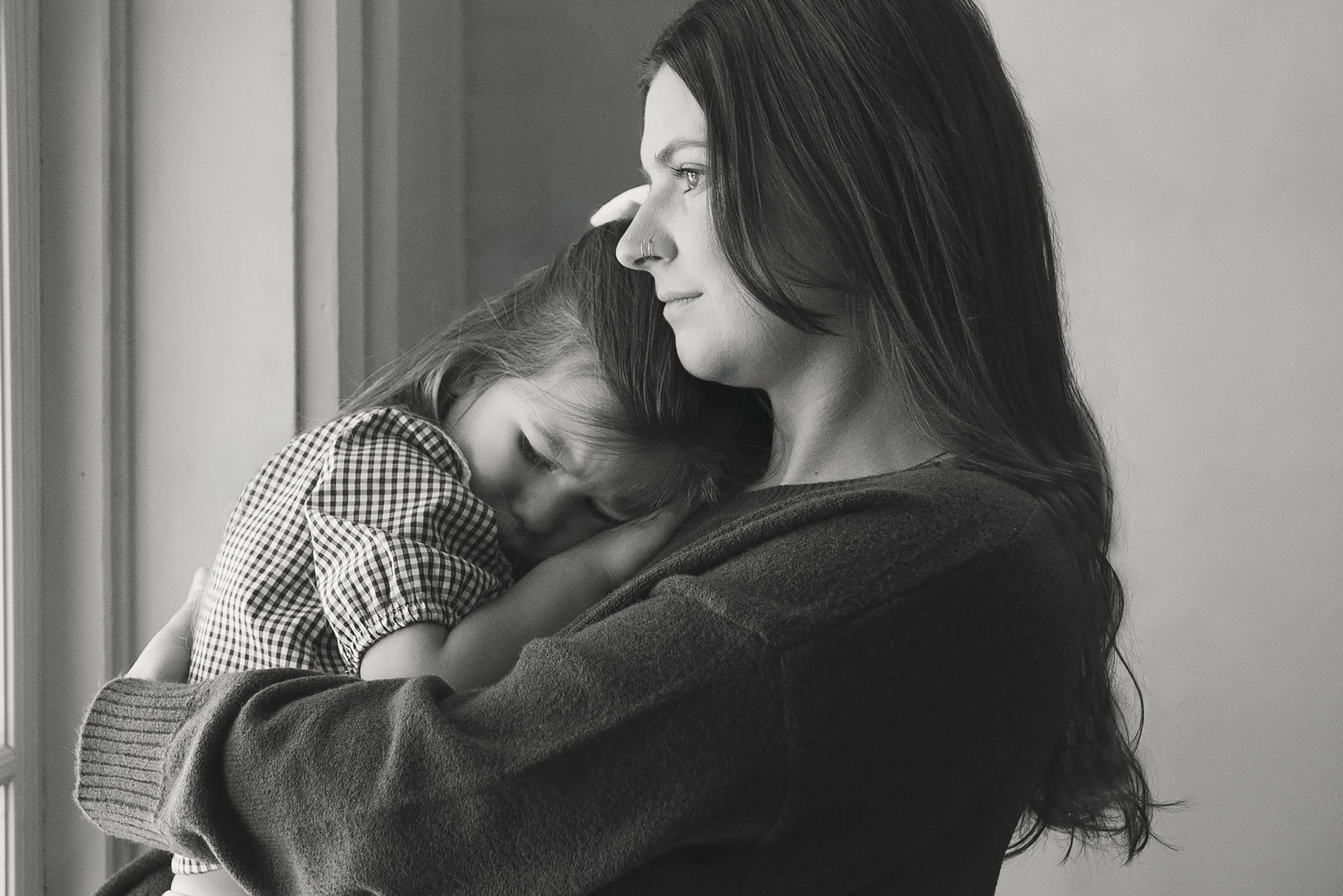 Black and white photo of a woman holding a small girl; the woman has long dark hair and a nose piercing, and the girl is resting her head on the woman's shoulder with her eyes closed.