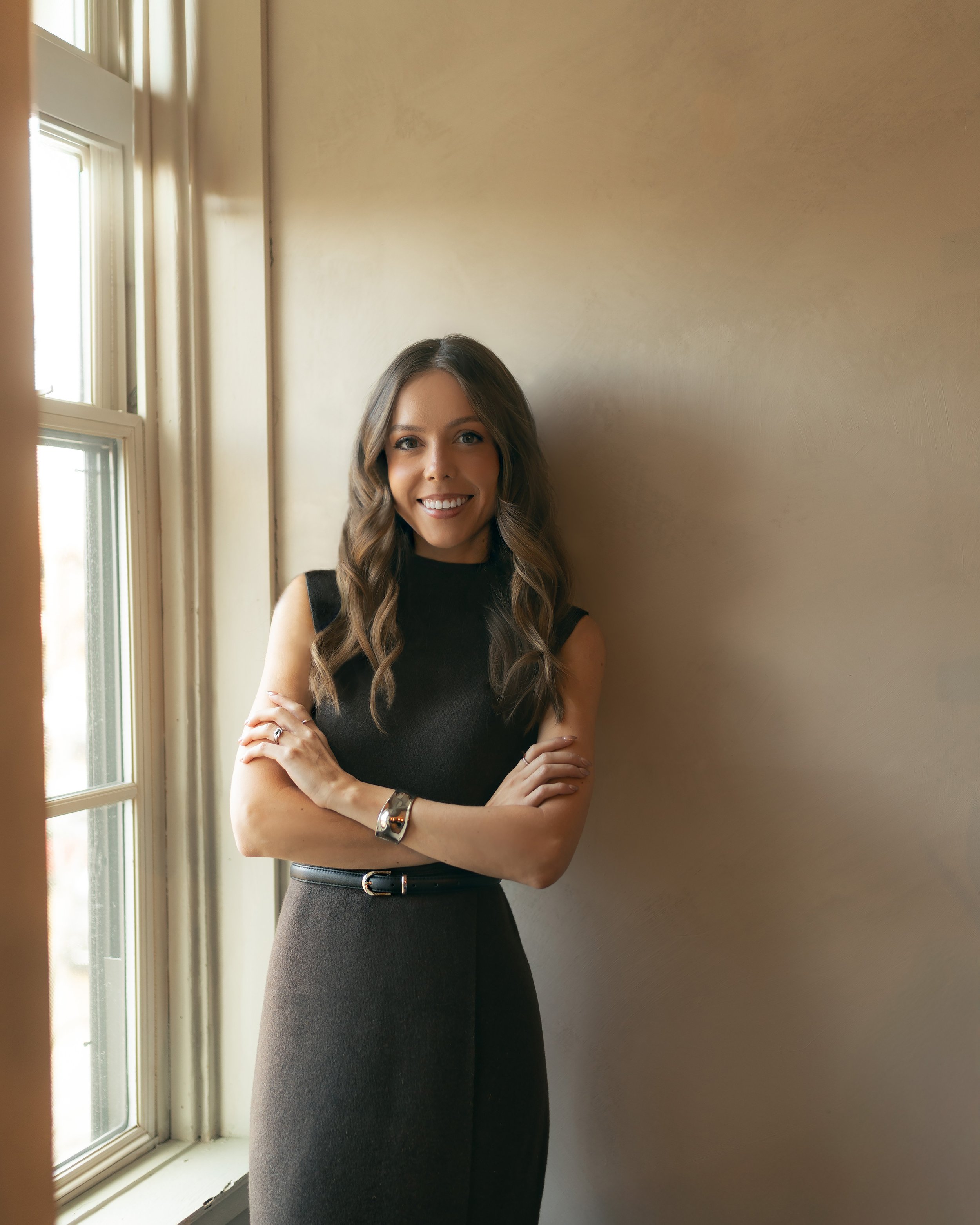 A woman with long wavy brown hair, wearing a sleeveless black top and a high-waisted dark skirt, stands near a window with her arms crossed and is smiling at the camera.
