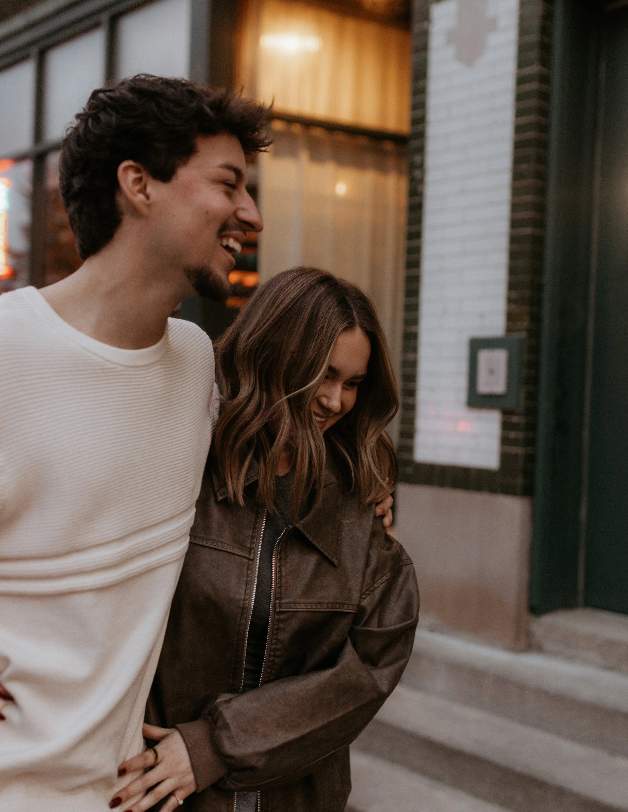 A young man and woman smiling and walking arm in arm outside a building at night.