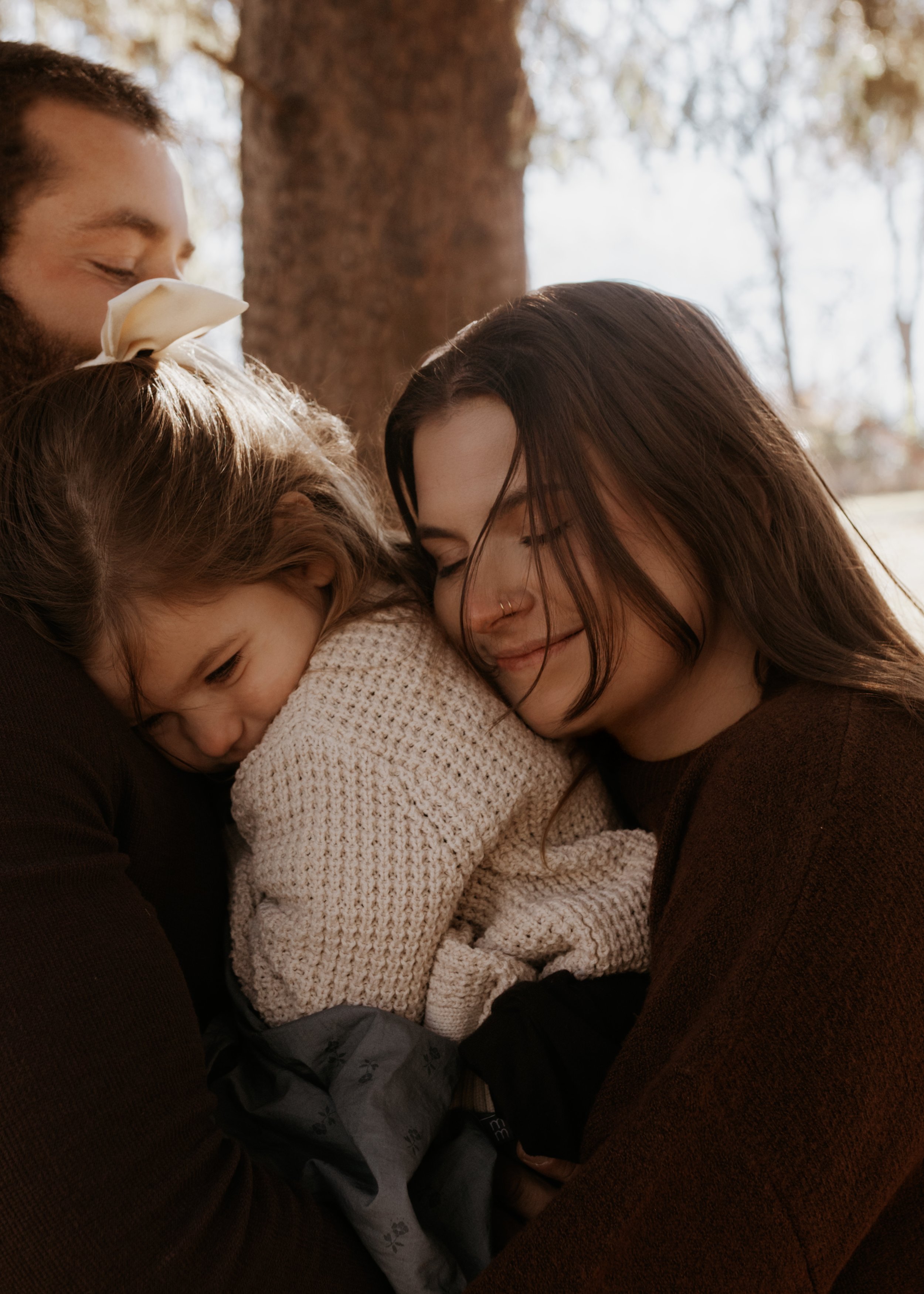 A family of four sharing a warm group hug outdoors, with trees in the background.