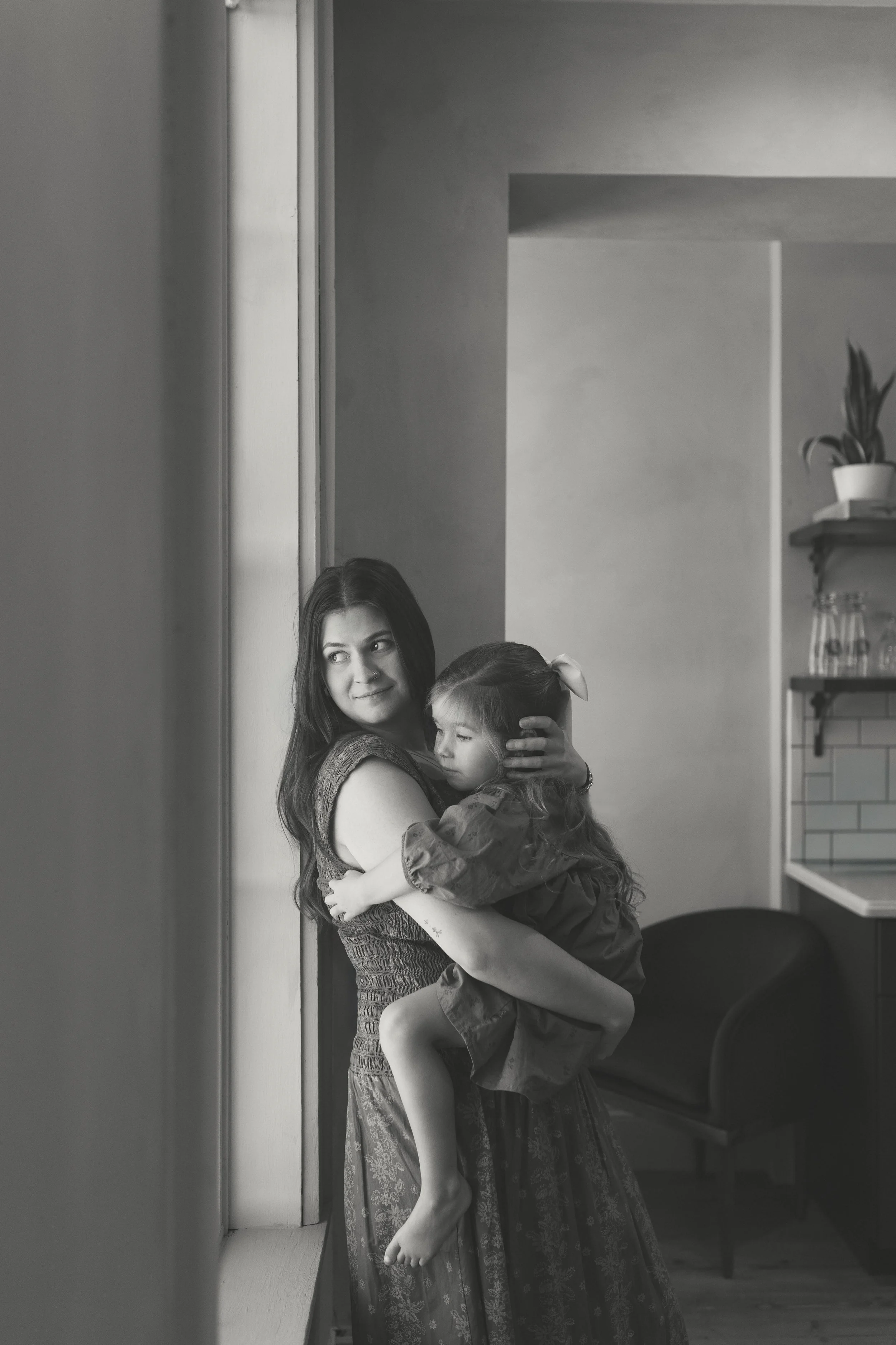 A woman holding a young girl close by a window in a home, with furniture and plants visible in the background.
