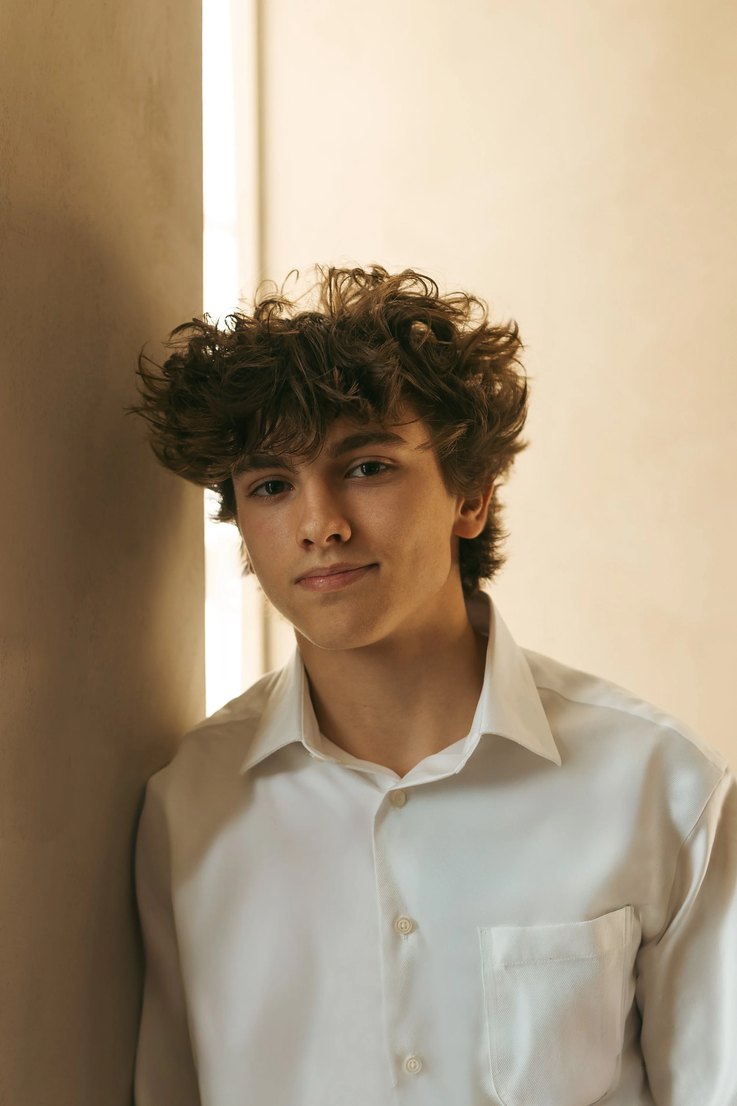 A young man with curly hair wearing a white dress shirt, standing against a wall with a light-colored background.