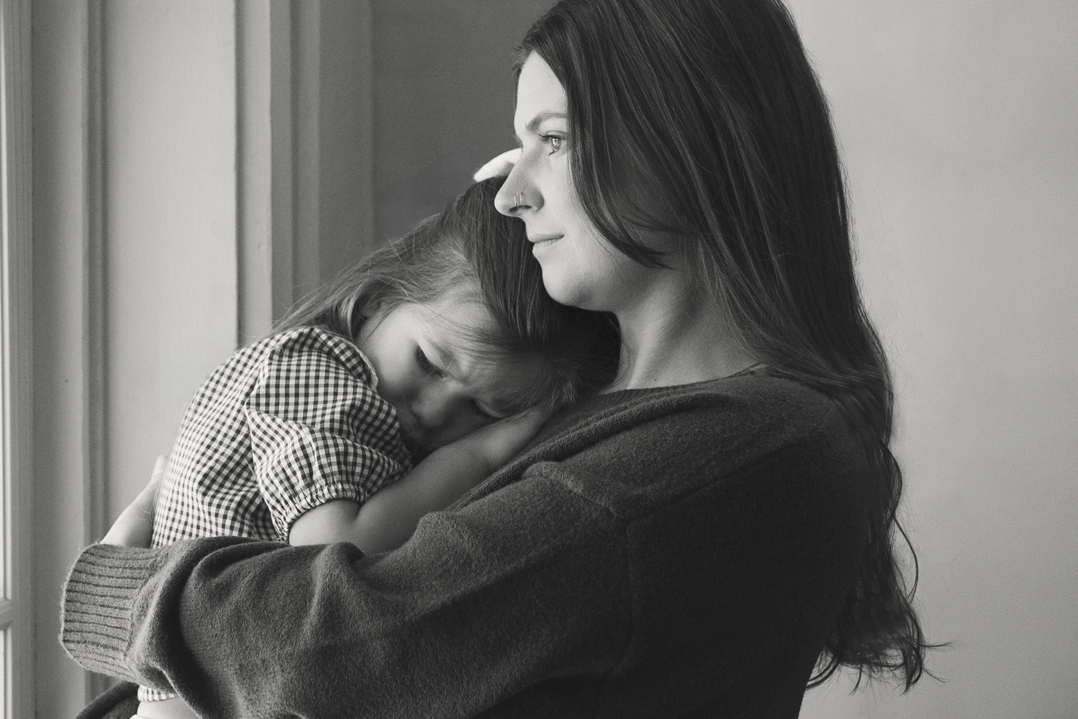 A woman holding a young girl, who is resting her head on her shoulder, near a window in black and white.