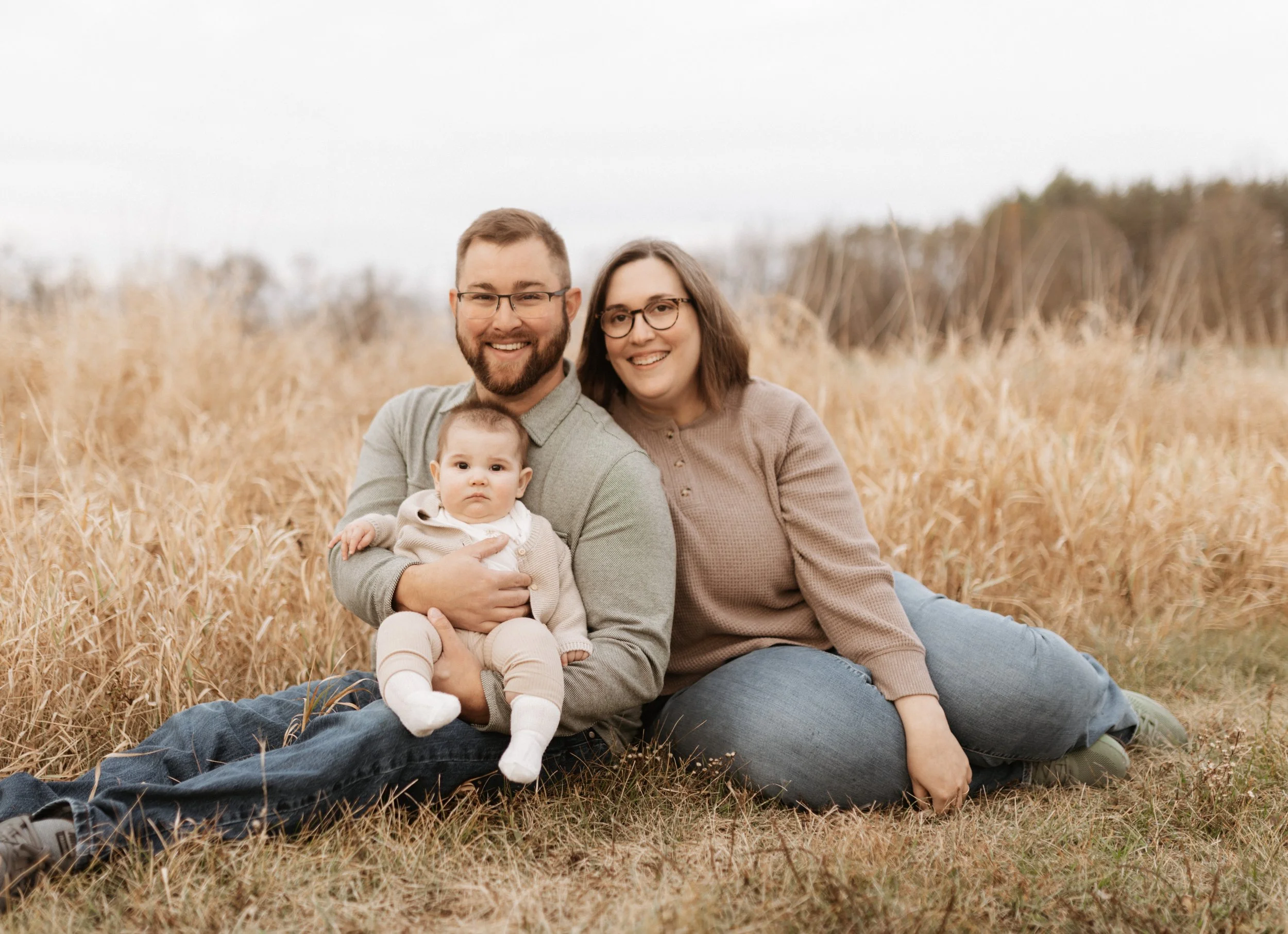 Family of three sitting and lying on the grass in a field of tall, golden grass, with an overcast sky in the background. The man has a beard and glasses, holding a baby girl. The woman has glasses and shoulder-length hair. All are smiling, dressed in
