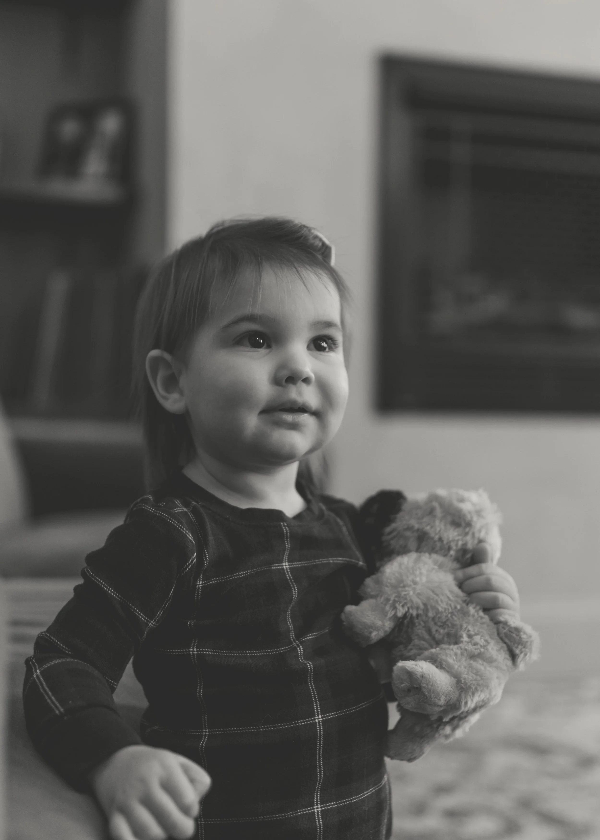 A young girl with medium-length hair holding a teddy bear, looking slightly upward in a room.