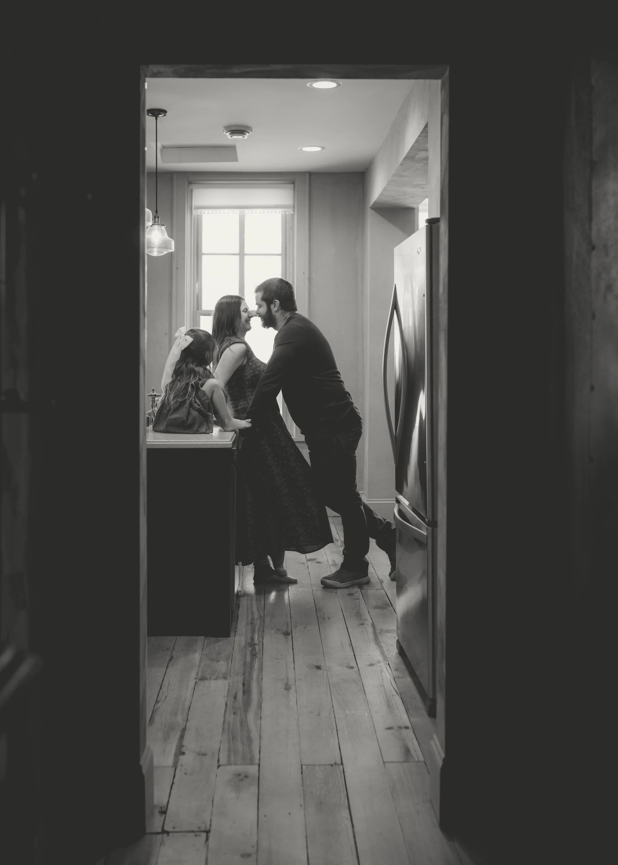 A black and white photo of a family in a kitchen through a doorway, showing a woman and a man leaning in close, smiling at each other, with a young girl nearby.