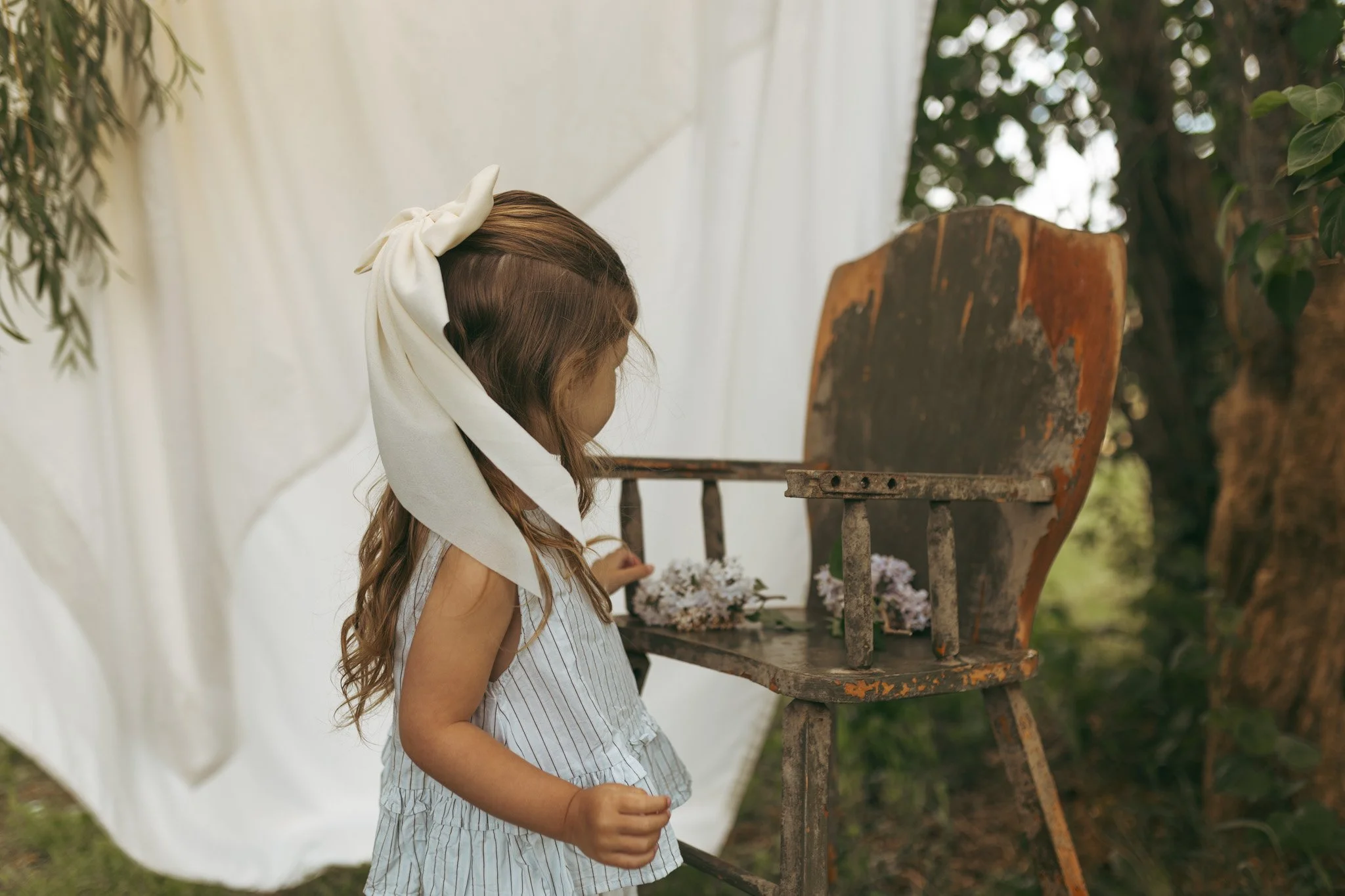 A young girl with long, wavy hair tied with a large white bow, wearing a sleeveless striped dress, standing outdoors near a rustic wooden chair with a white fabric backdrop and trees in the background.