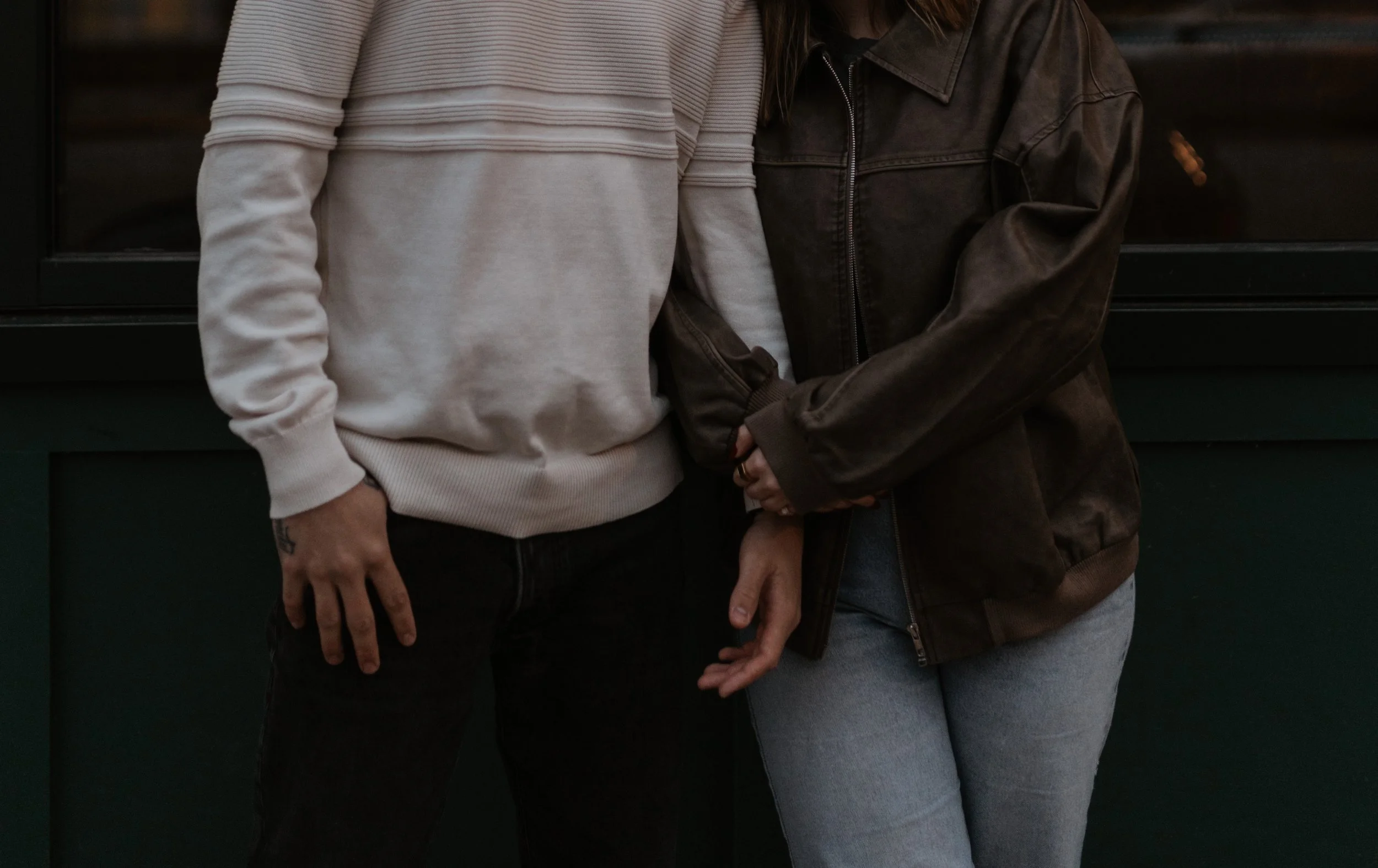 Close-up of two people standing together, one wearing a light-colored sweater and dark pants, the other in a dark jacket and light jeans, with their hands near each other.