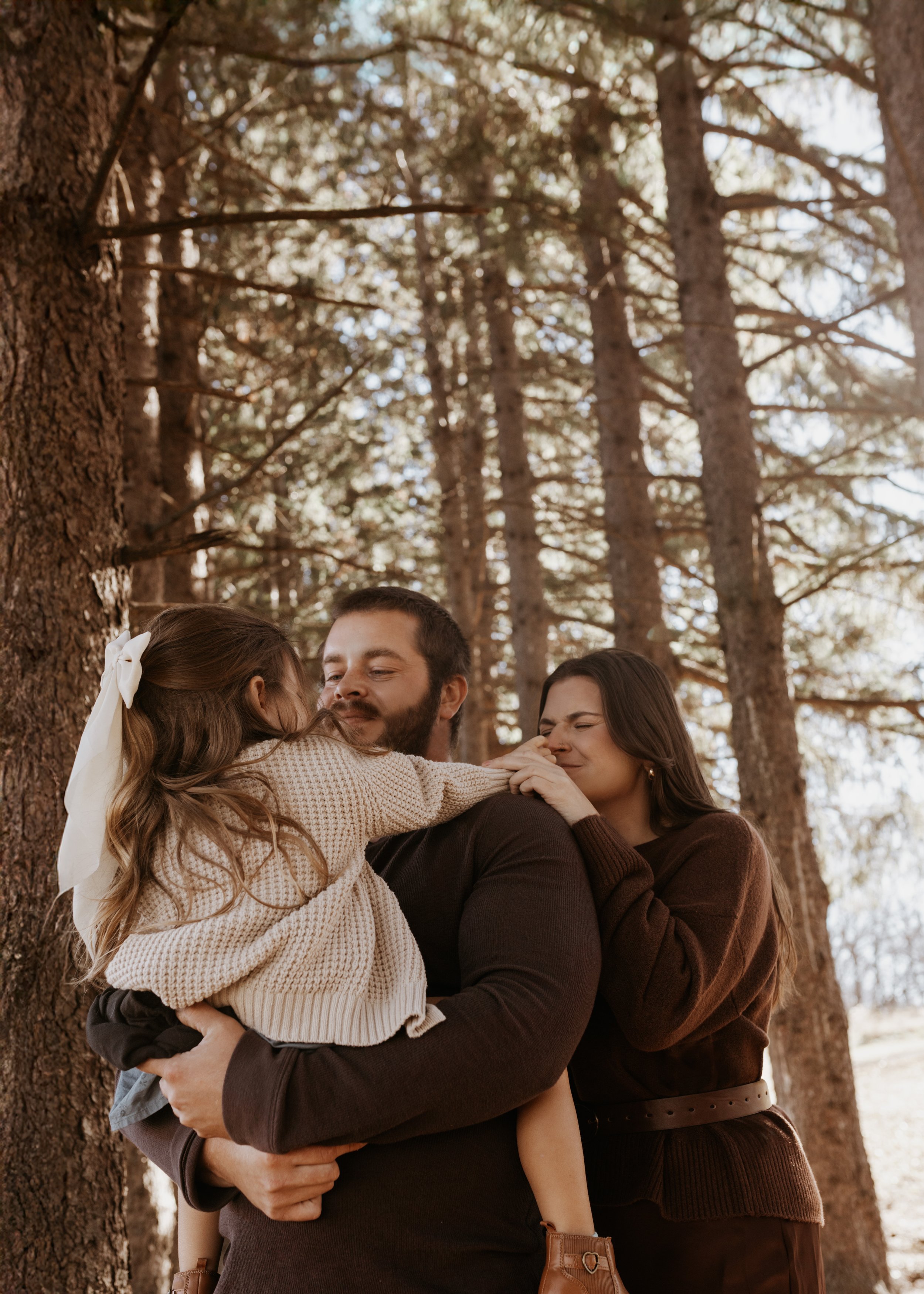 A man holding a girl in his arms while a woman touches his shoulder in a forest with tall trees.