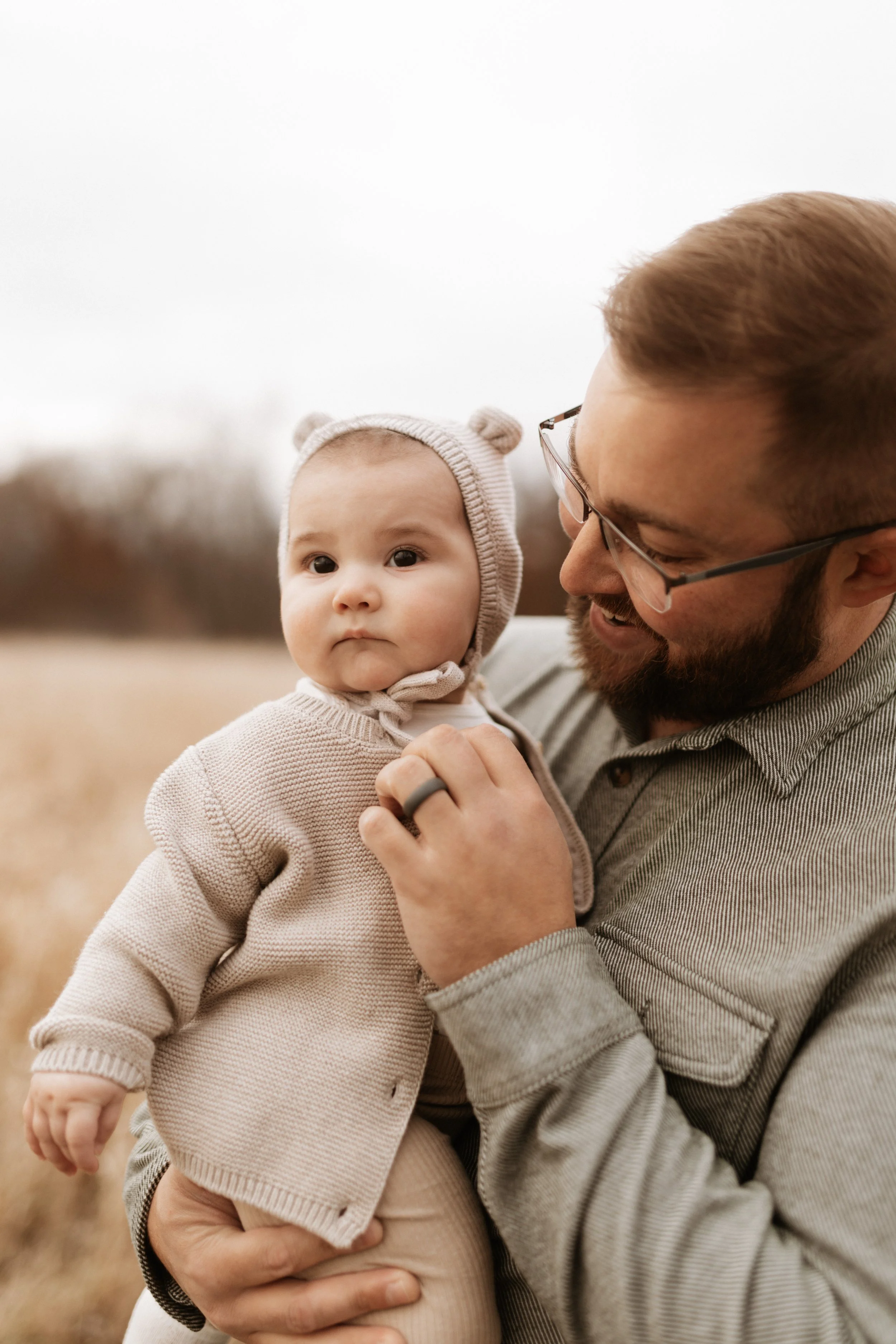 A man holding a baby girl outdoors in a field with overcast sky, the man wearing glasses and a striped shirt, the baby dressed in a beige sweater and a knit hat with bear ears.