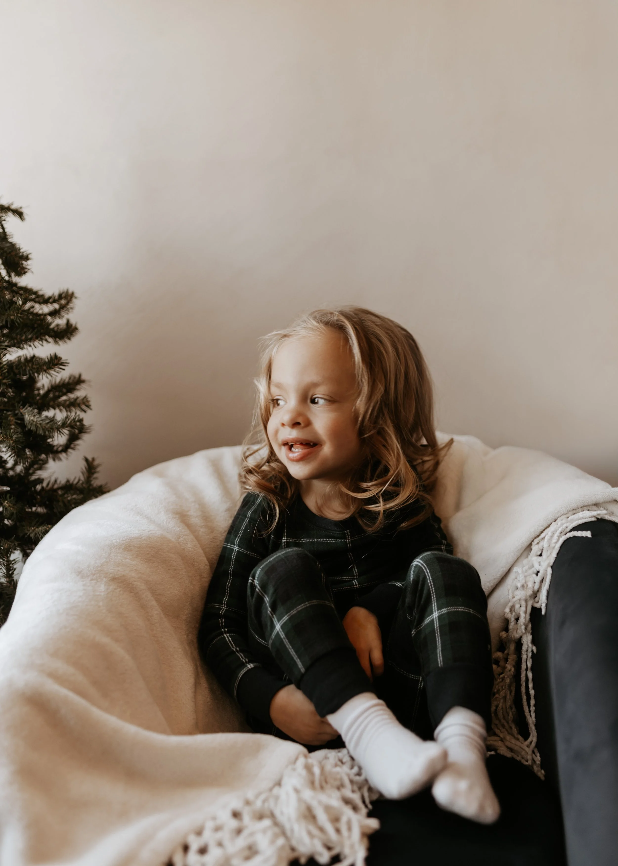 Young girl with light brown hair sitting on a cozy beige blanket on a black chair, smiling and looking to her left, with a Christmas tree partially visible on the left side.