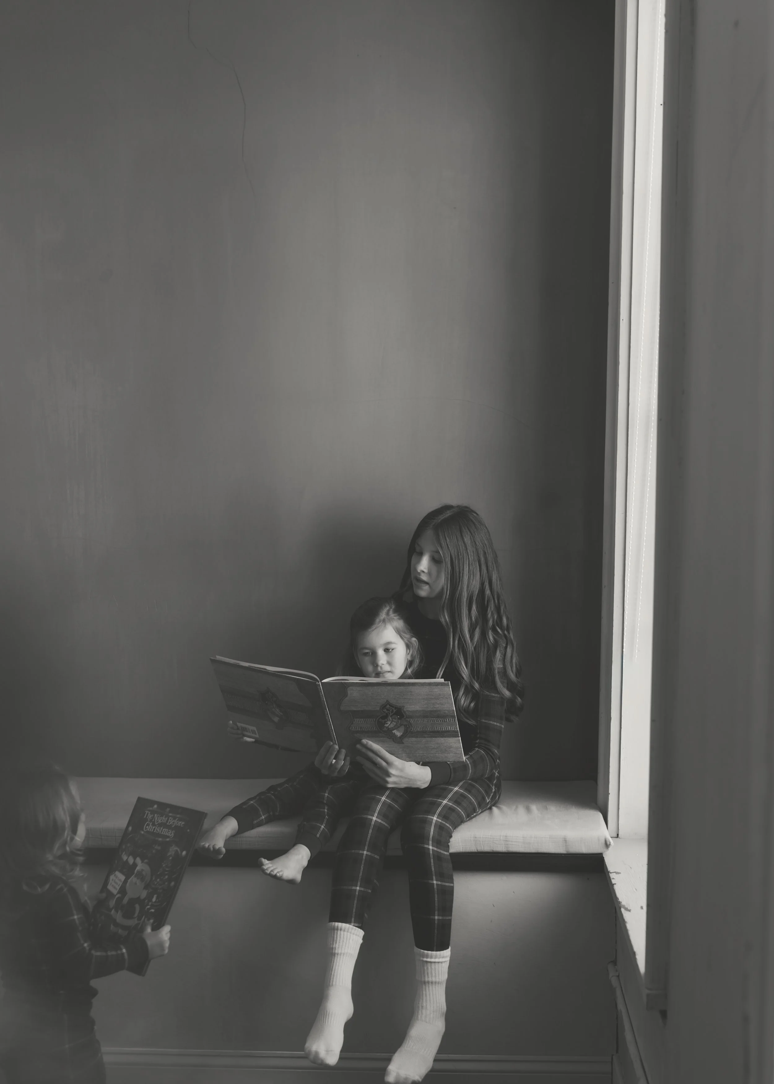 Two girls in pajamas are reading books together on a window seat in a room. One girl is sitting with her legs crossed, the other is sitting with her legs hanging over the edge. Another girl, also in pajamas, is standing in front of them holding a boo