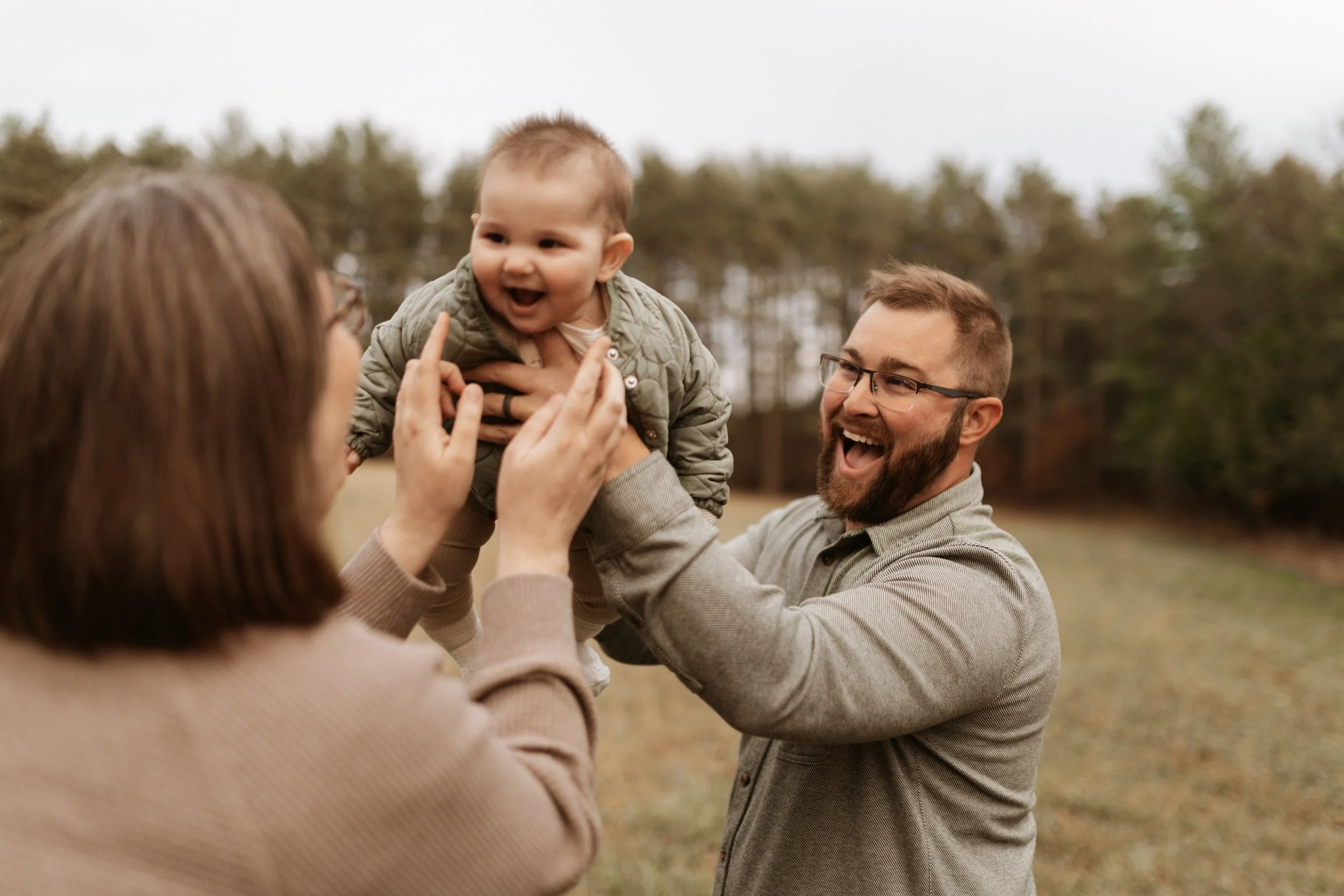 A man with glasses and a woman are smiling as they hold a joyful baby boy in an outdoor park with trees in the background.