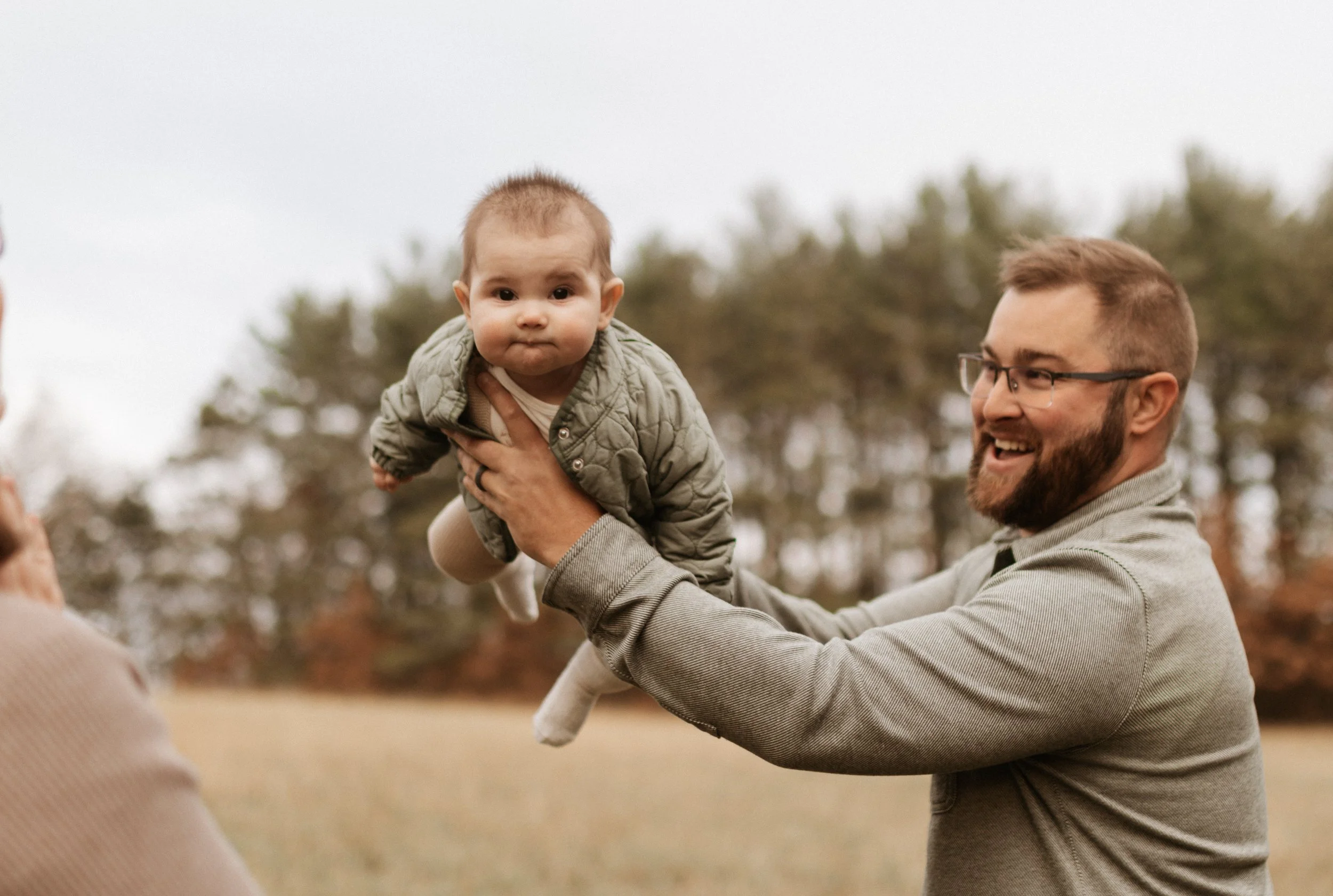 A man with glasses and a beard is holding a young child in the air outdoors in a field with trees in the background. The man is smiling, and the child has a curious expression.