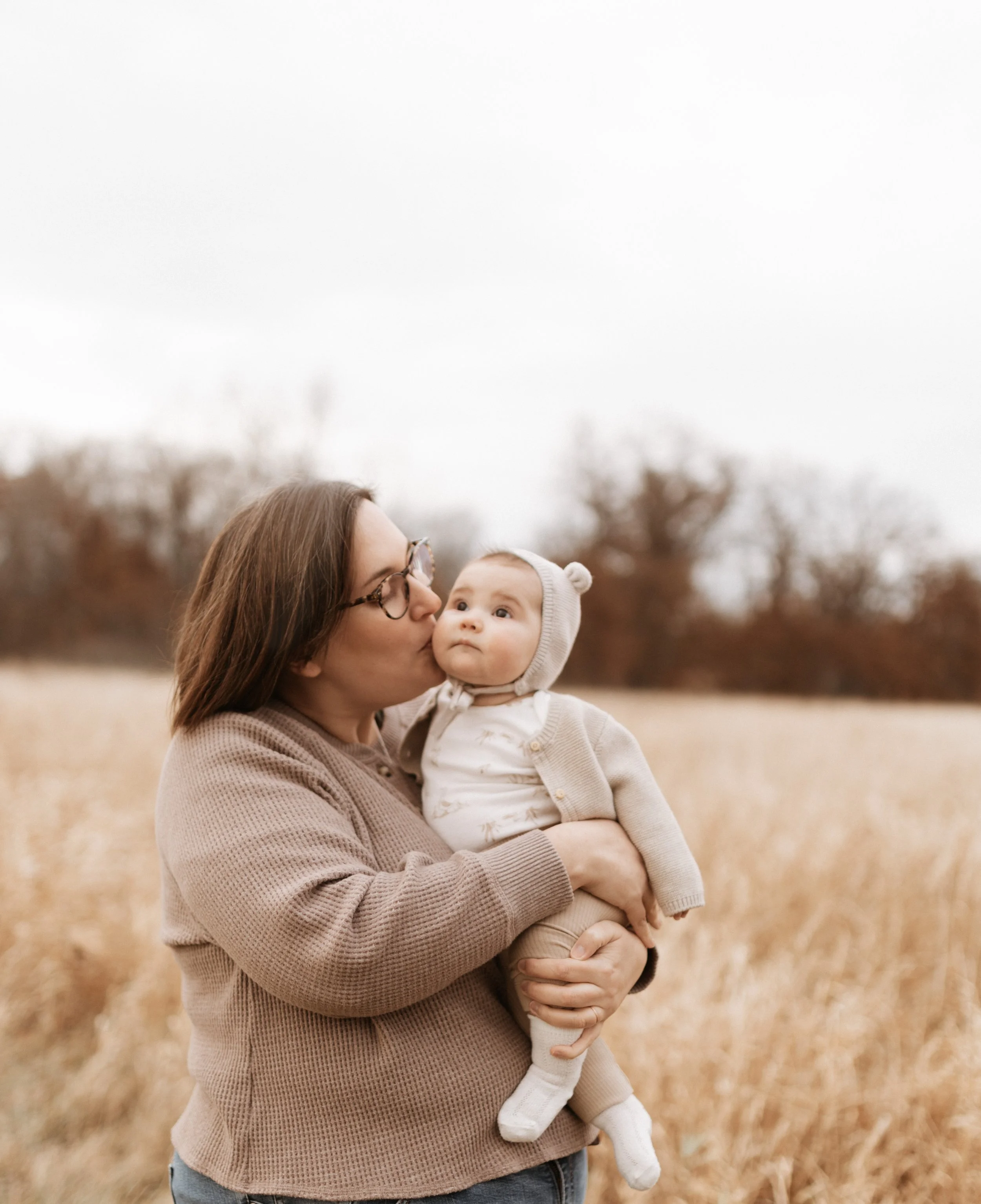 A woman holding a baby outdoors in a field of tall, golden grass, with trees in the background and an overcast sky.