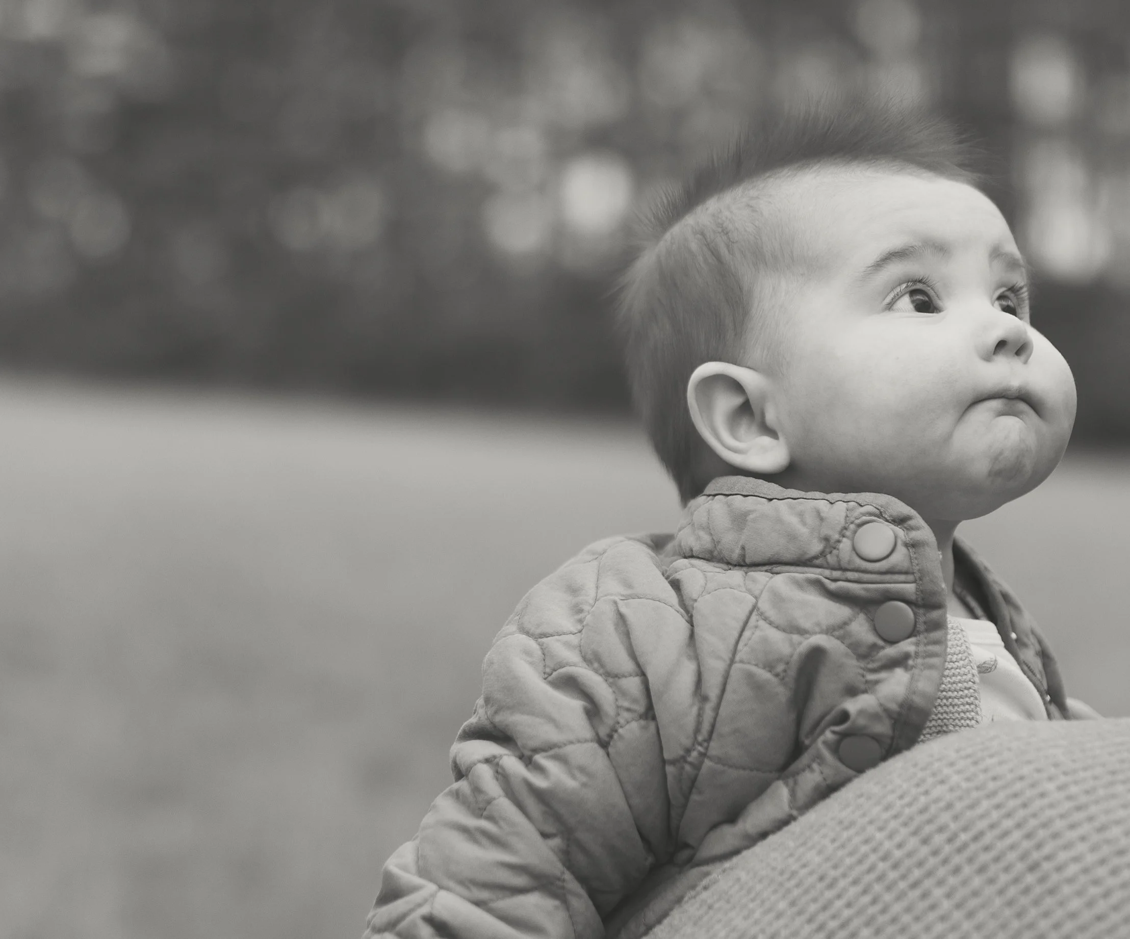 A black-and-white photo of a young child with a round face, short hair, and expressive eyes, sitting outdoors and looking upwards.