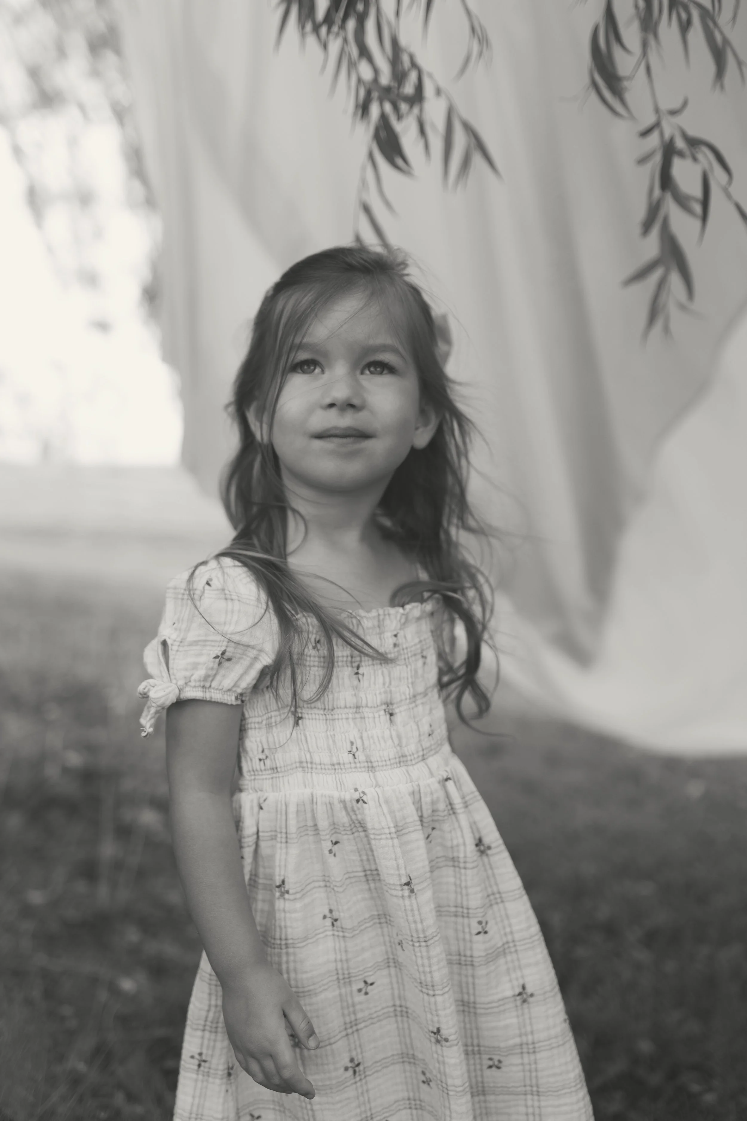 A young girl with long hair standing outdoors near a curtain of hanging fabric, with trees in the background.