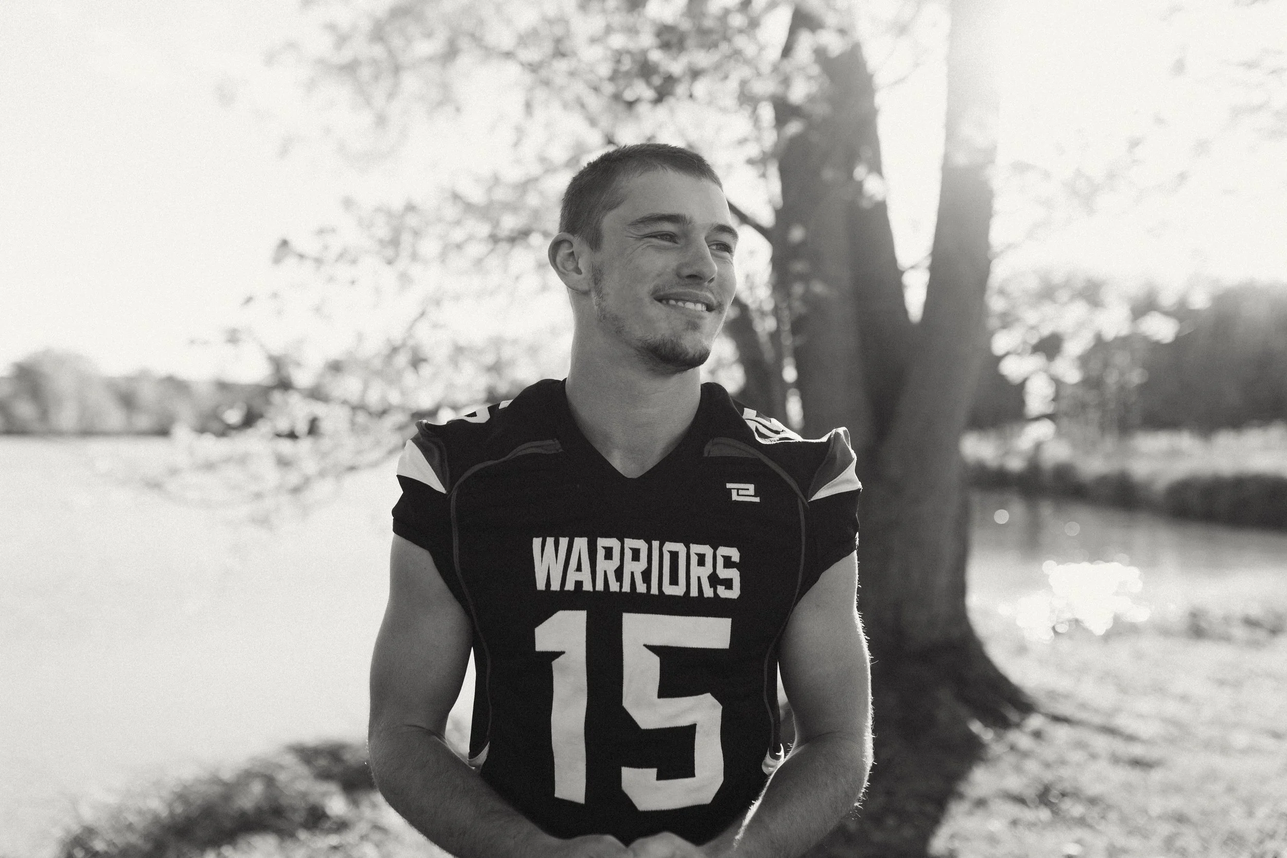 Young man in football uniform with number 15 and 'Warriors' on the chest, smiling outdoors near a tree by a body of water during sunset.
