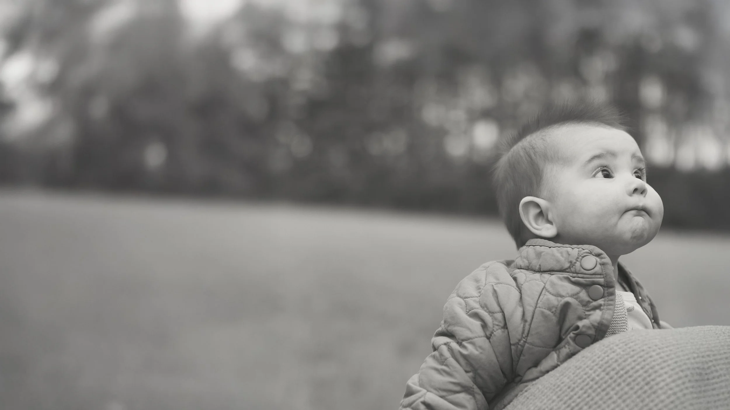Black and white photo of a young child with a mohawk hairstyle, wearing a quilted jacket, looking upwards with a puffed-cheek expression, outdoors in a park or open space with trees in the background.