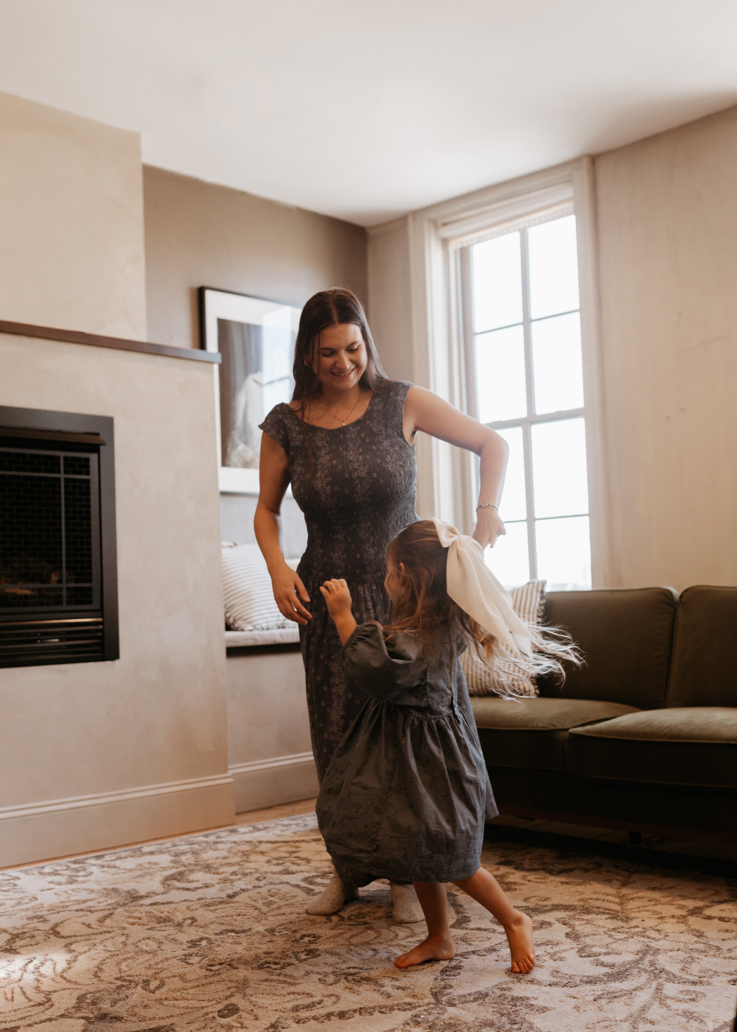 A woman and a young girl dancing indoors in a living room with a patterned rug, a couch, a window with sunlight, and a fireplace.