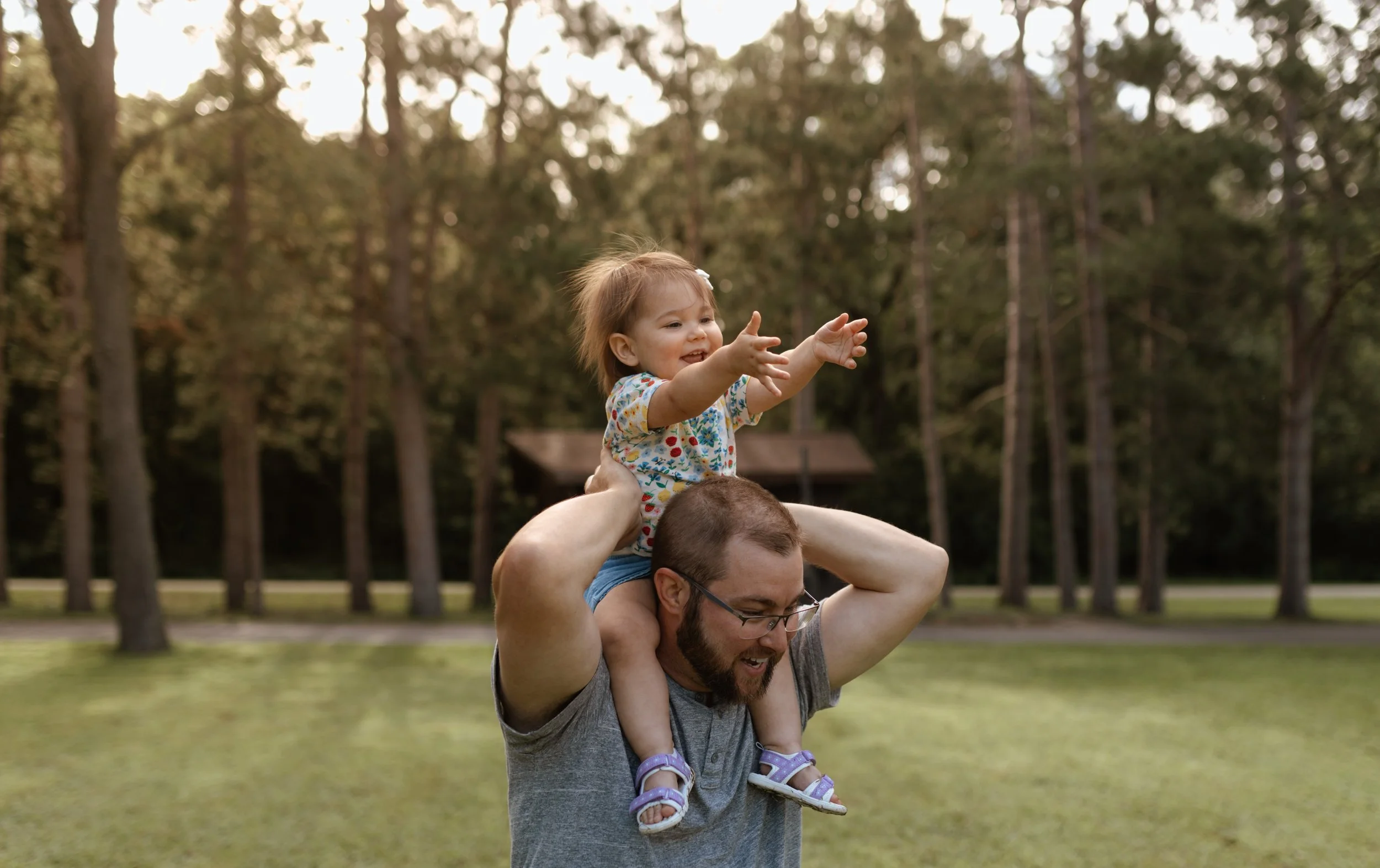A man with glasses is carrying a young girl on his shoulders in a park with trees in the background. The girl is reaching out with her hands and smiling.