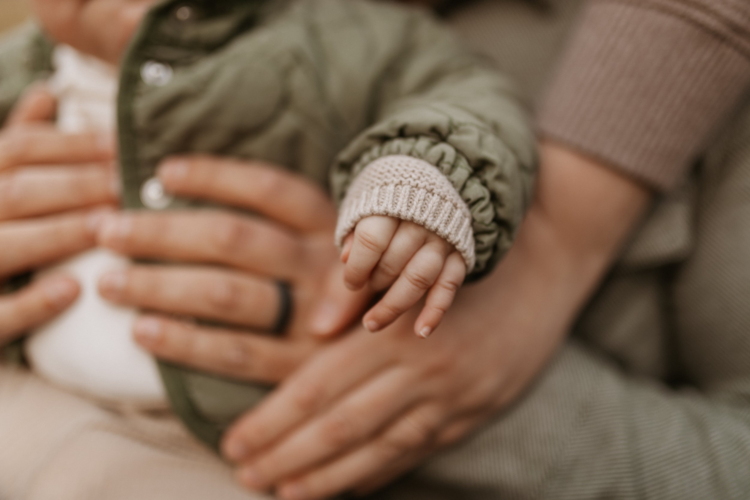 Close-up of a child's tiny hand holding an adult's hand, with both hands in focus. The child is wearing a beige knitted sleeve and the adult is wearing a ring and has a darker skin tone.