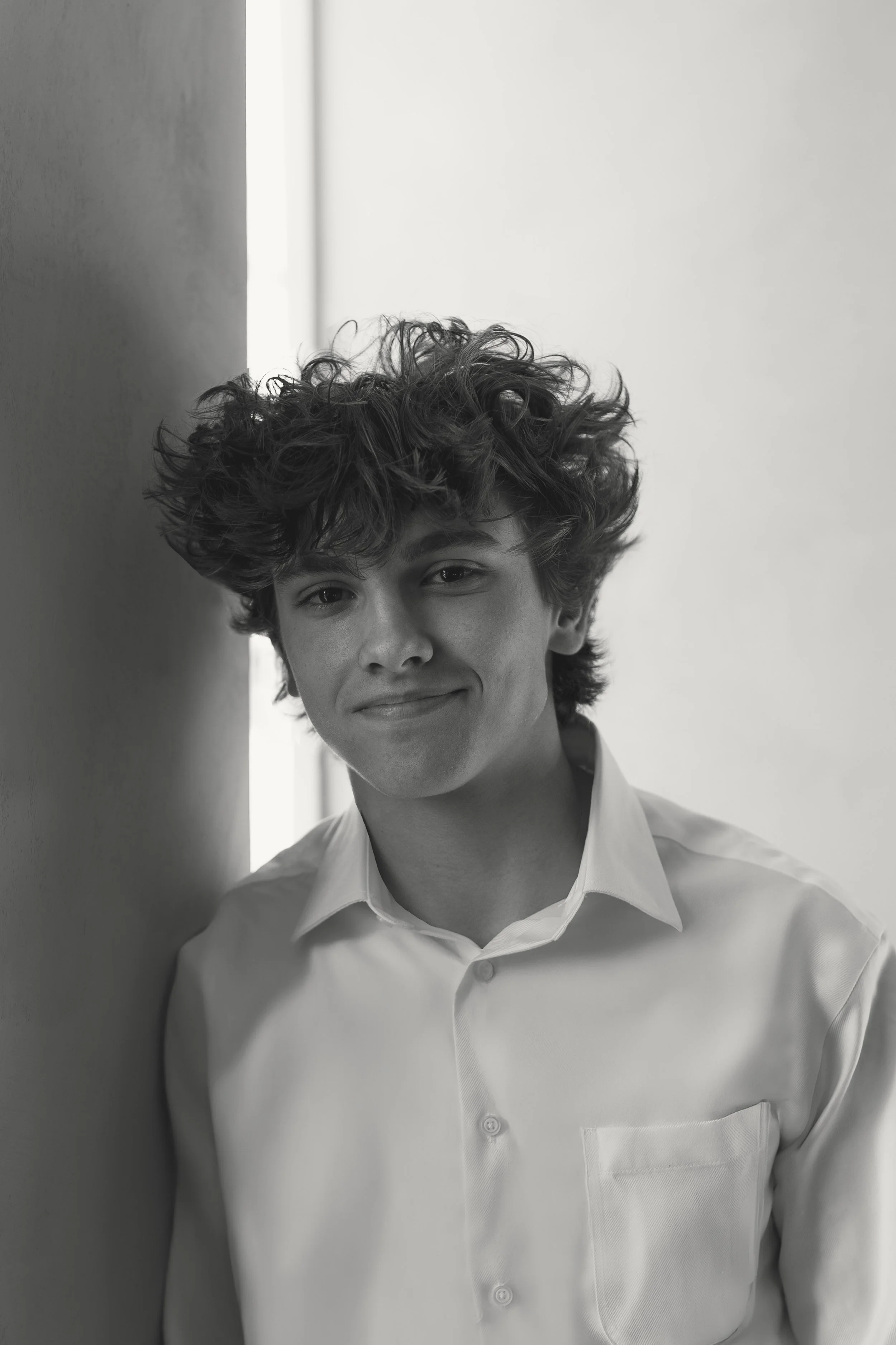 A young man with curly hair and a slight smile, wearing a white shirt, leaning against a wall in a black-and-white portrait.