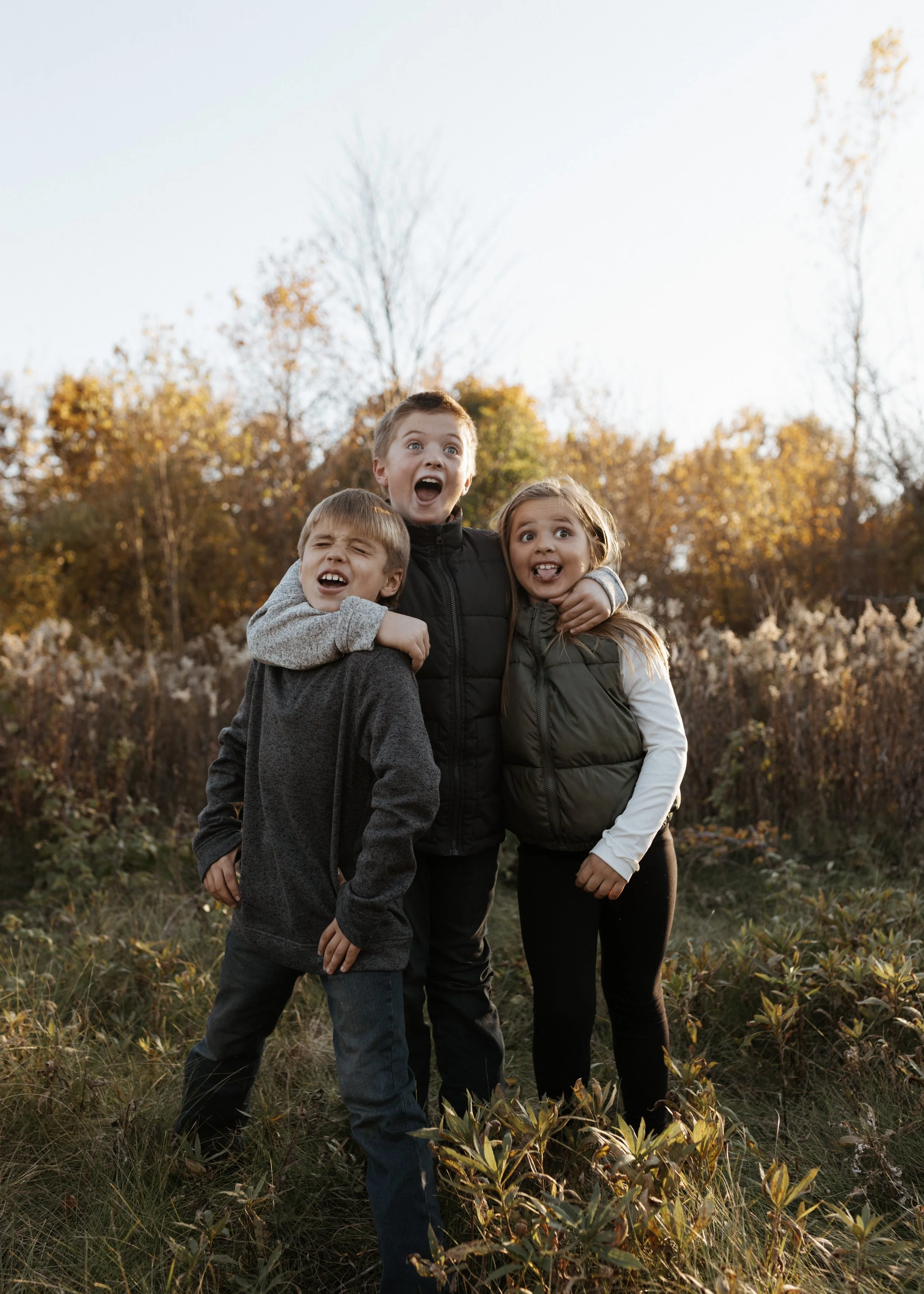 Three children outdoors in a field during autumn, with trees and golden foliage in the background, making funny faces and posing closely together.
