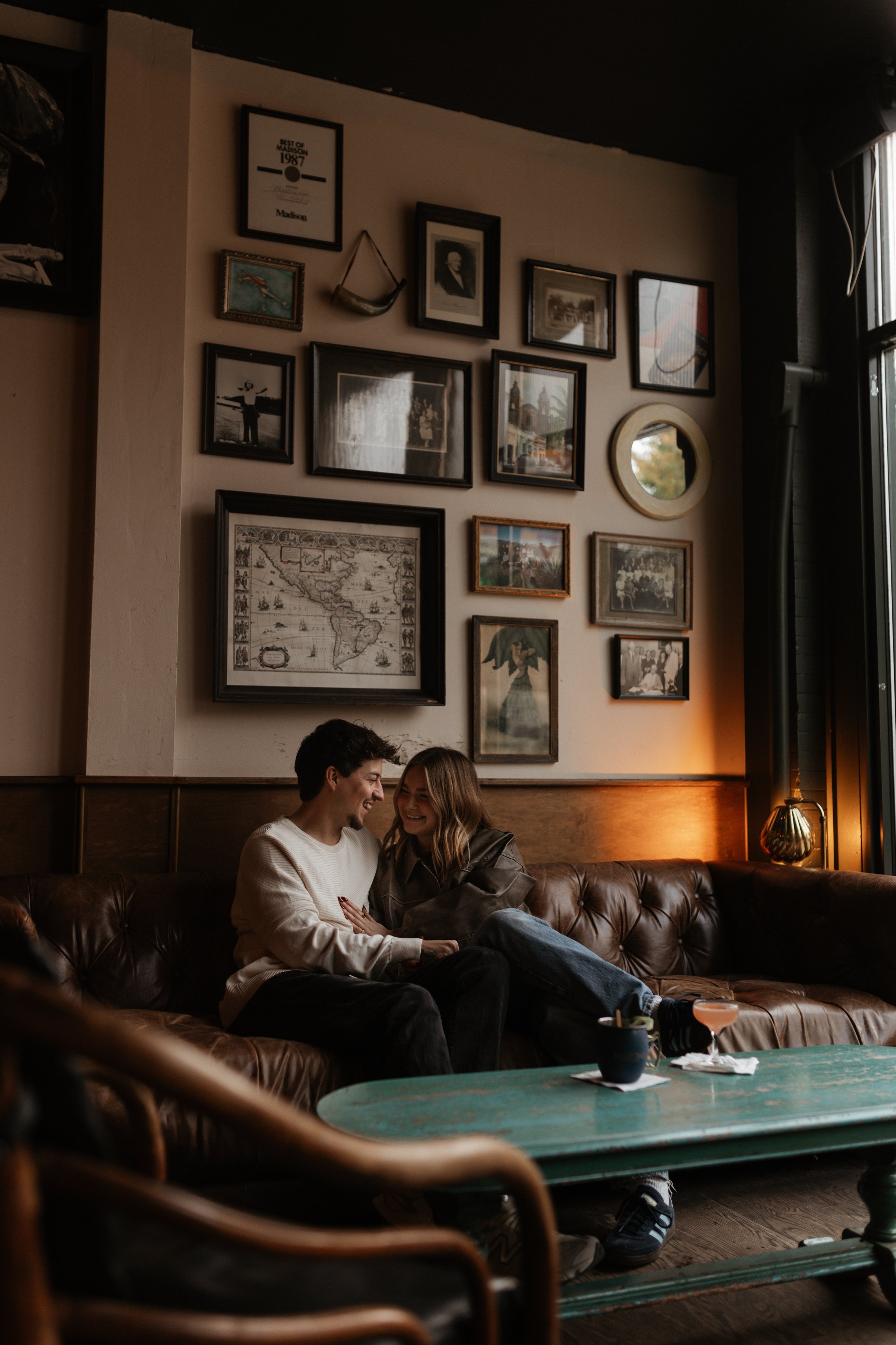 A young couple sitting on a brown leather couch in a cozy cafe, smiling and laughing together.