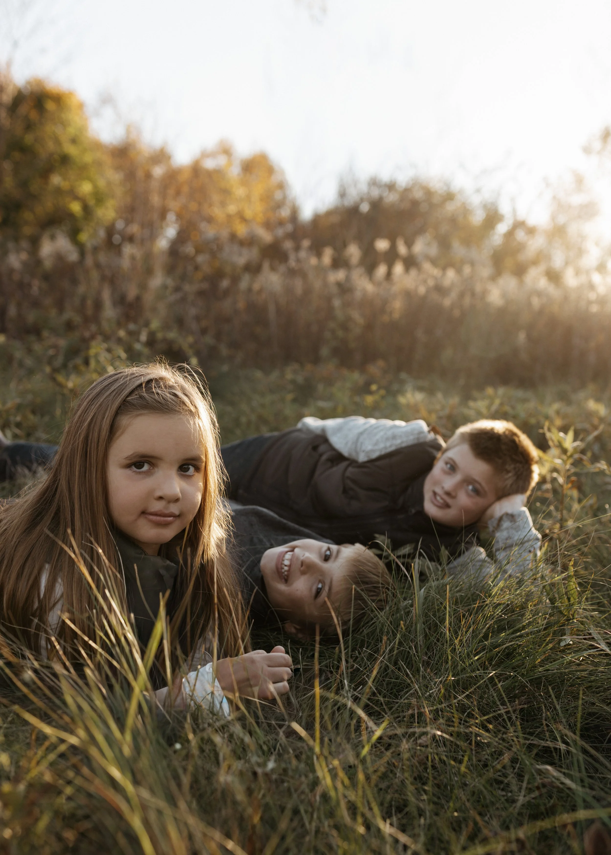Three children lying in tall grass outdoors during late afternoon, with trees and sunlight in the background.