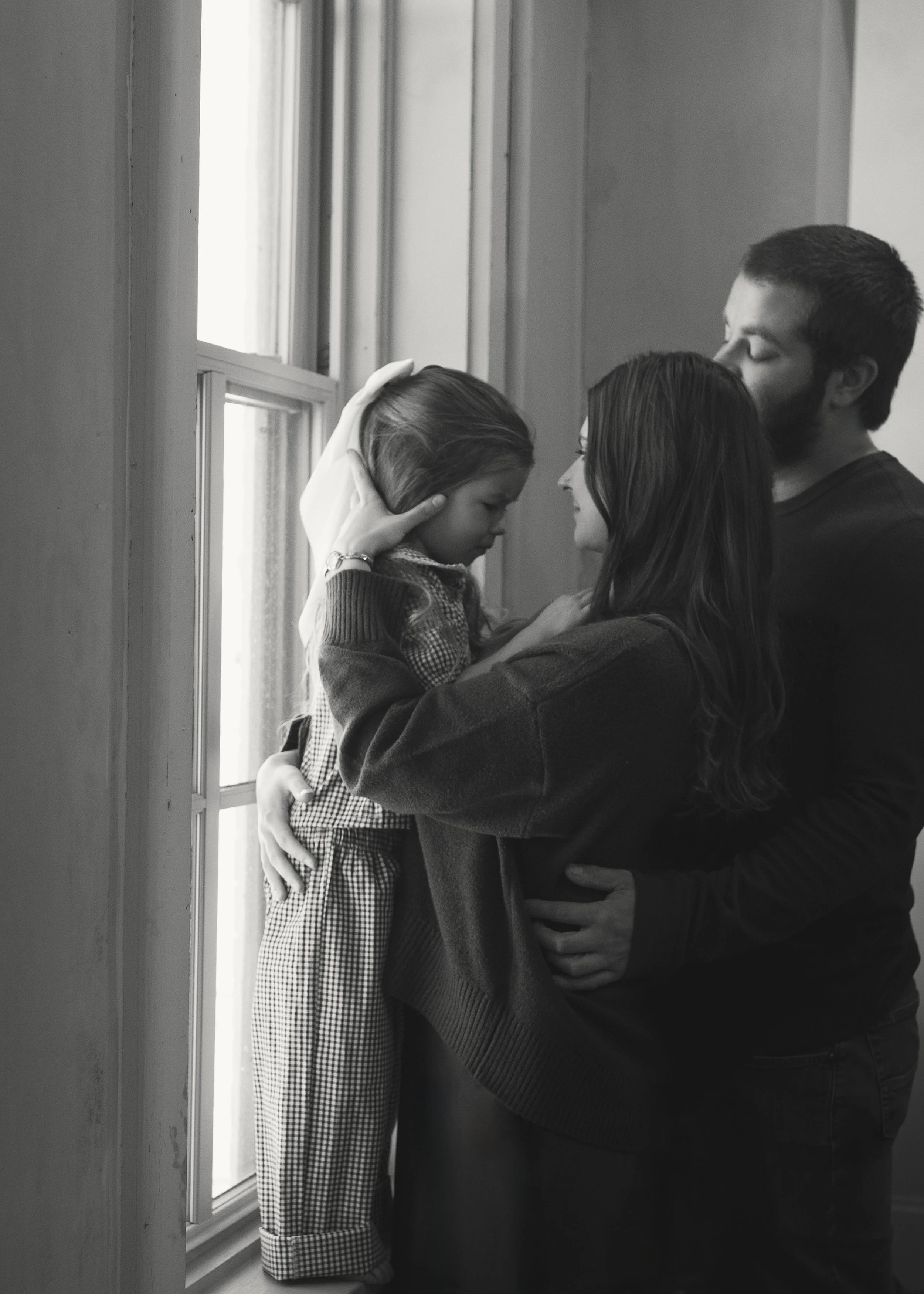 A family of three standing by a window, with the mother and father gently comforting their young daughter by holding her face and waist.