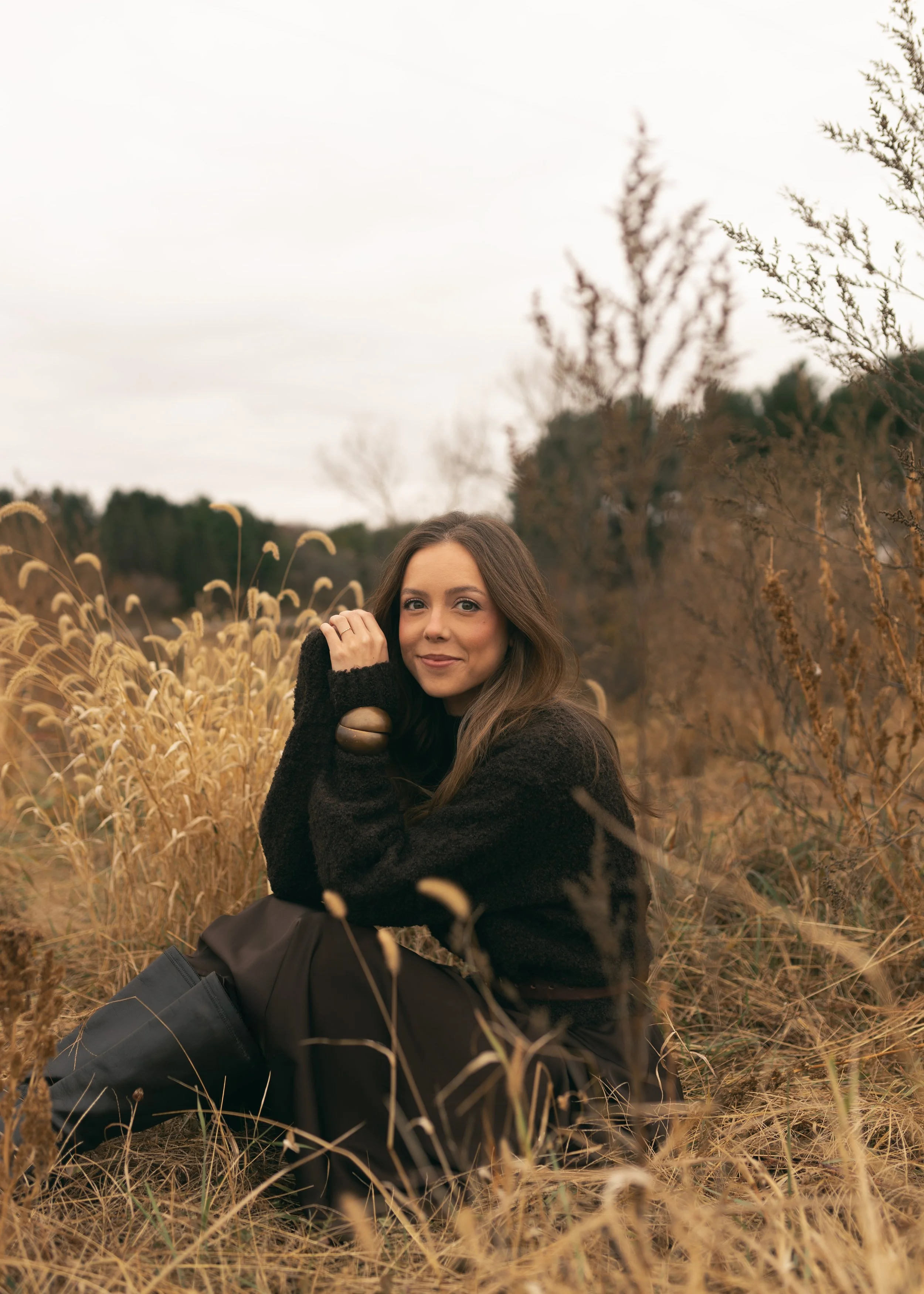 A woman sitting in a field of tall, dry grass with trees in the background on a cloudy day, smiling at the camera.