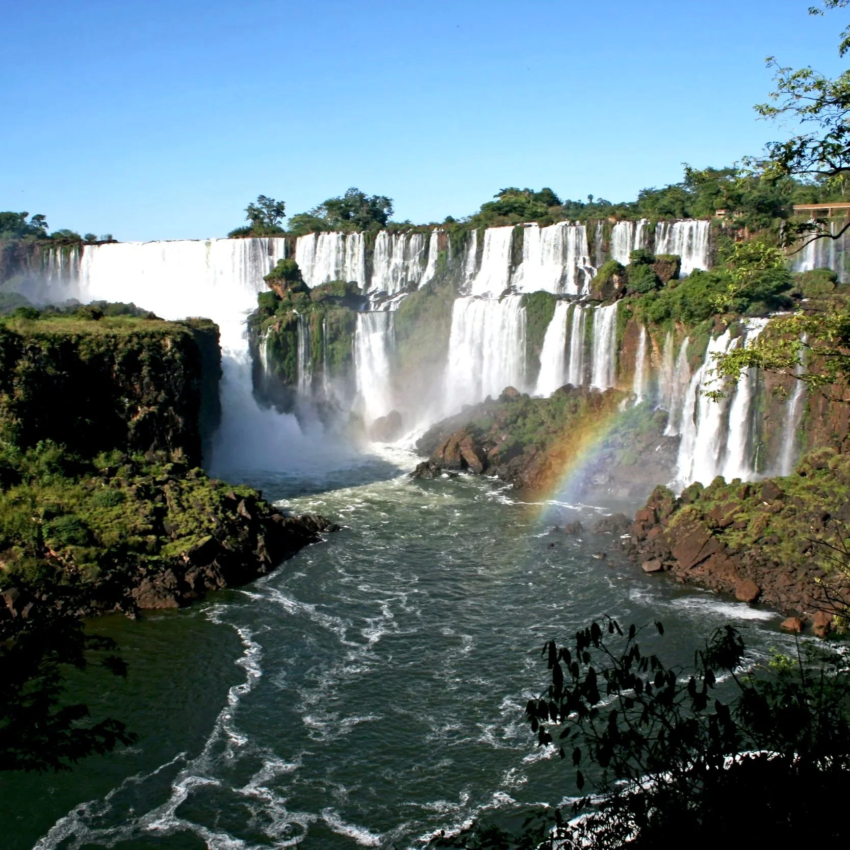 View of Iguazu Falls with multiple waterfalls, lush greenery on the riverbanks, a rainbow near the base of the falls, and a clear blue sky.