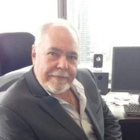 A woman with shoulder-length black hair, smiling outdoors in front of green leafy plants, wearing a blue top.