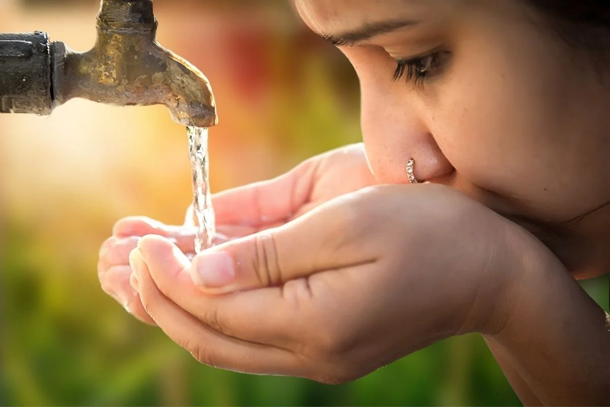 A woman drinking water from her hands from an outdoor faucet during sunset.