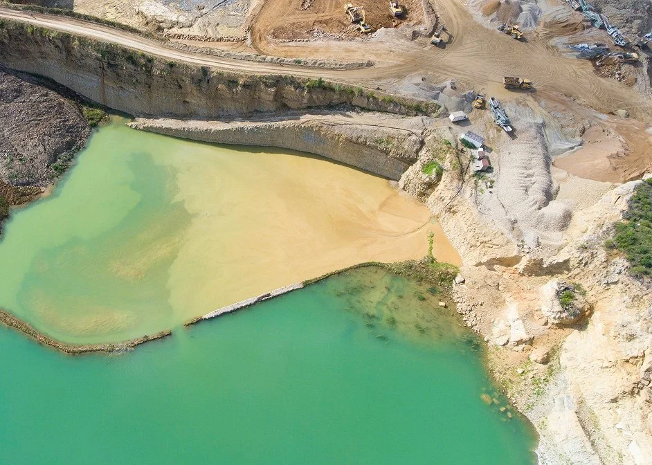 Aerial view of a landscape with a pond divided into two sections, one greenish and one yellowish, surrounded by rocky terrain and earthworks.
