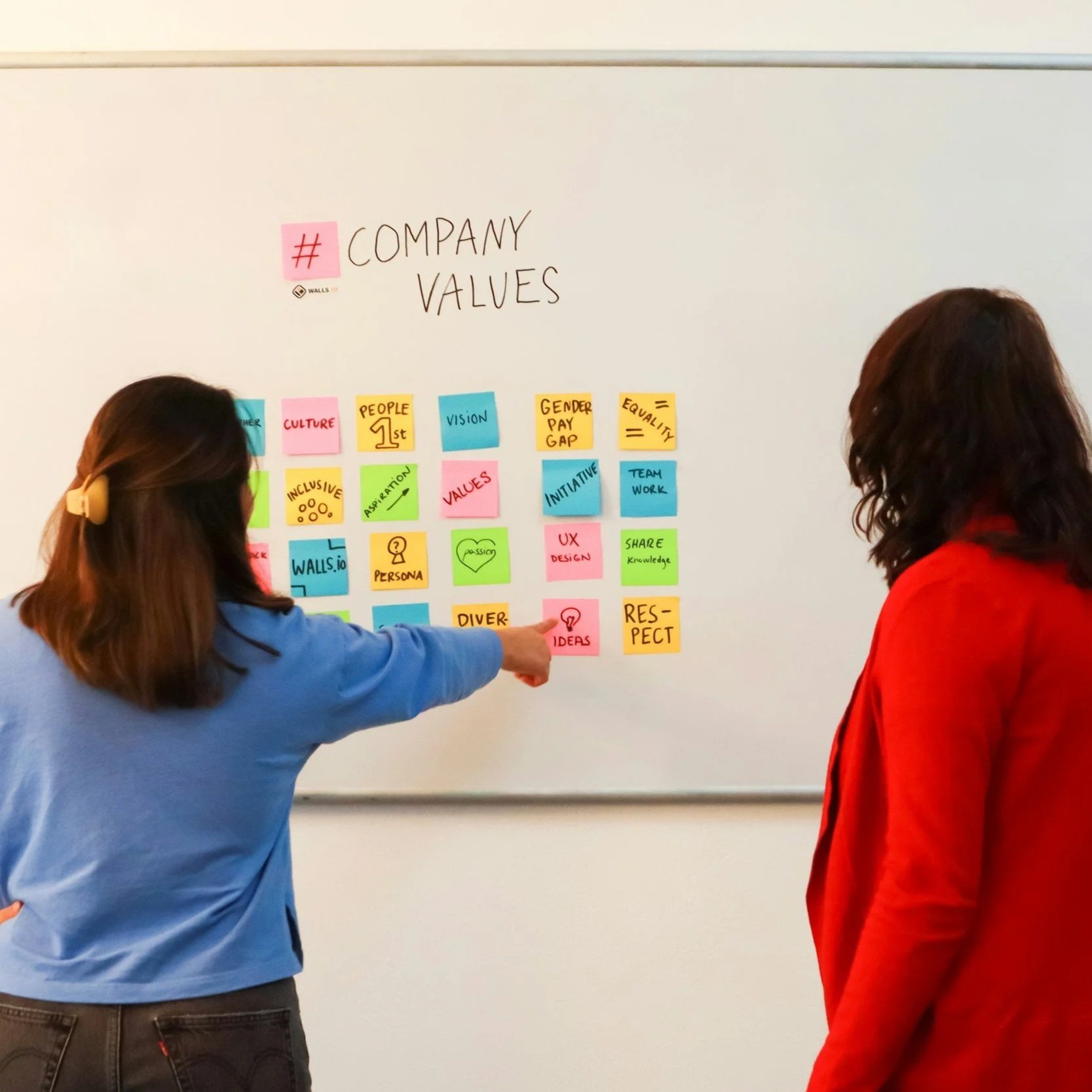Two women discussing company values written on sticky notes on a whiteboard.