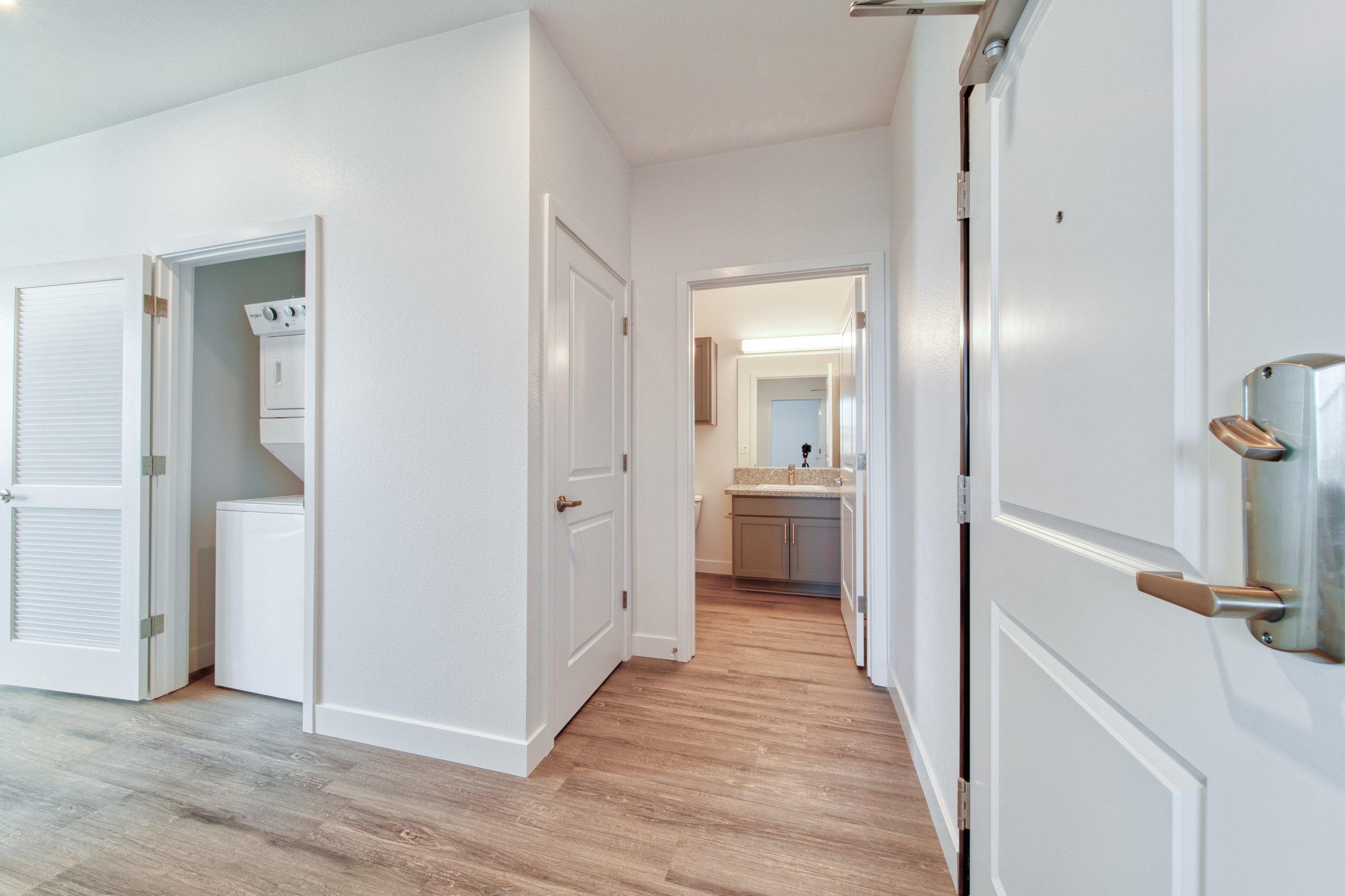 Interior view of an apartment or house showing a hallway with a laundry closet, bathroom with vanity and mirror, and hardwood flooring.