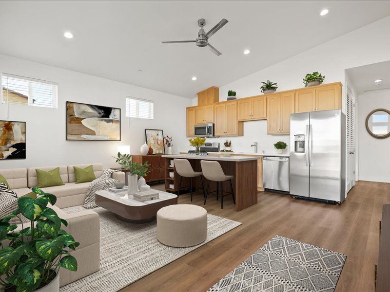 Open-concept living room and kitchen with modern decor, beige sofa, green pillows, abstract artwork, wooden cabinetry, stainless steel appliances, kitchen island with barstools, and potted plants.