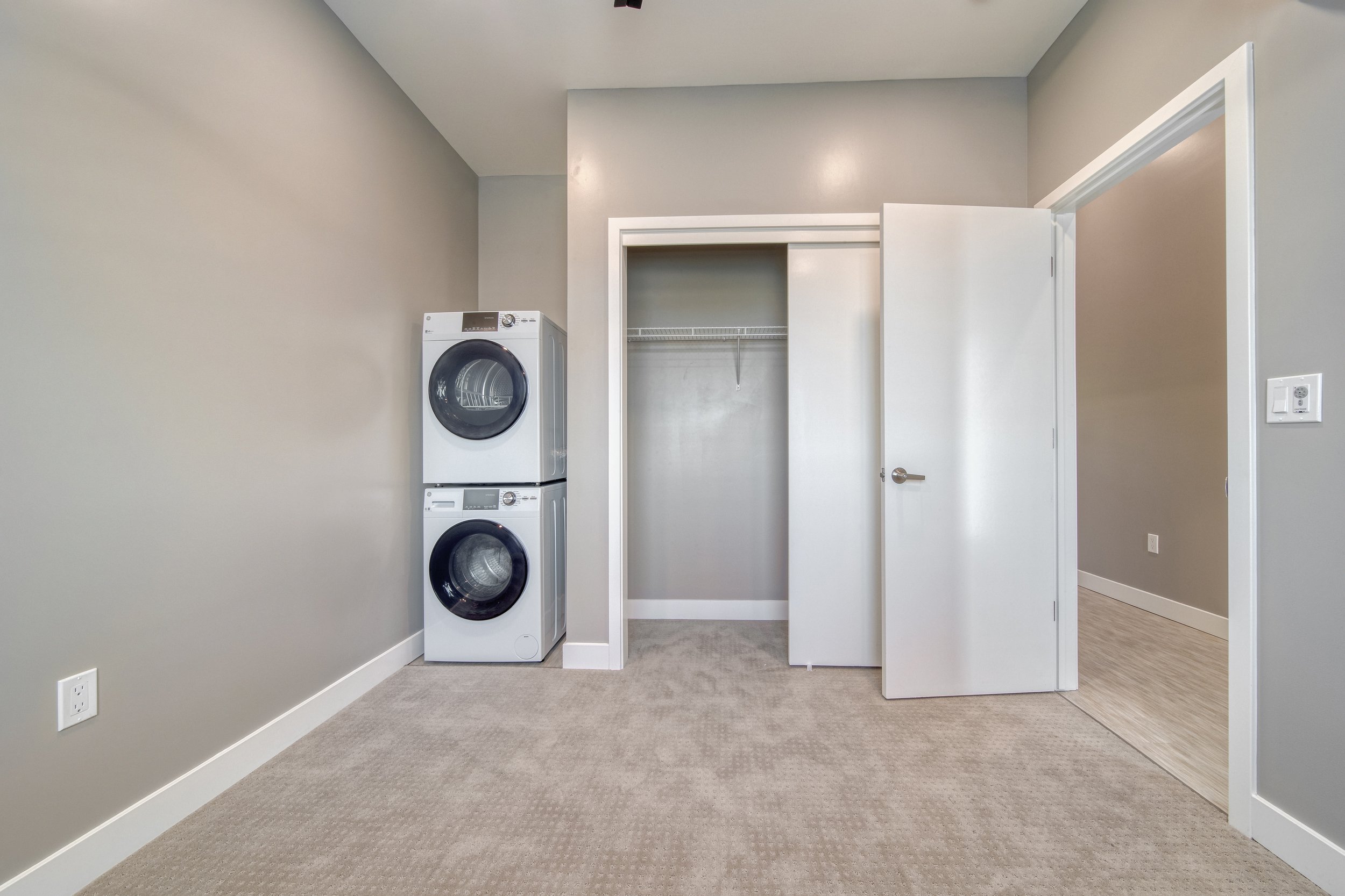 Empty laundry room with stacked washer and dryer, open closet with wire shelf, beige carpet, gray walls, white door, light-colored laminate flooring in adjacent room.