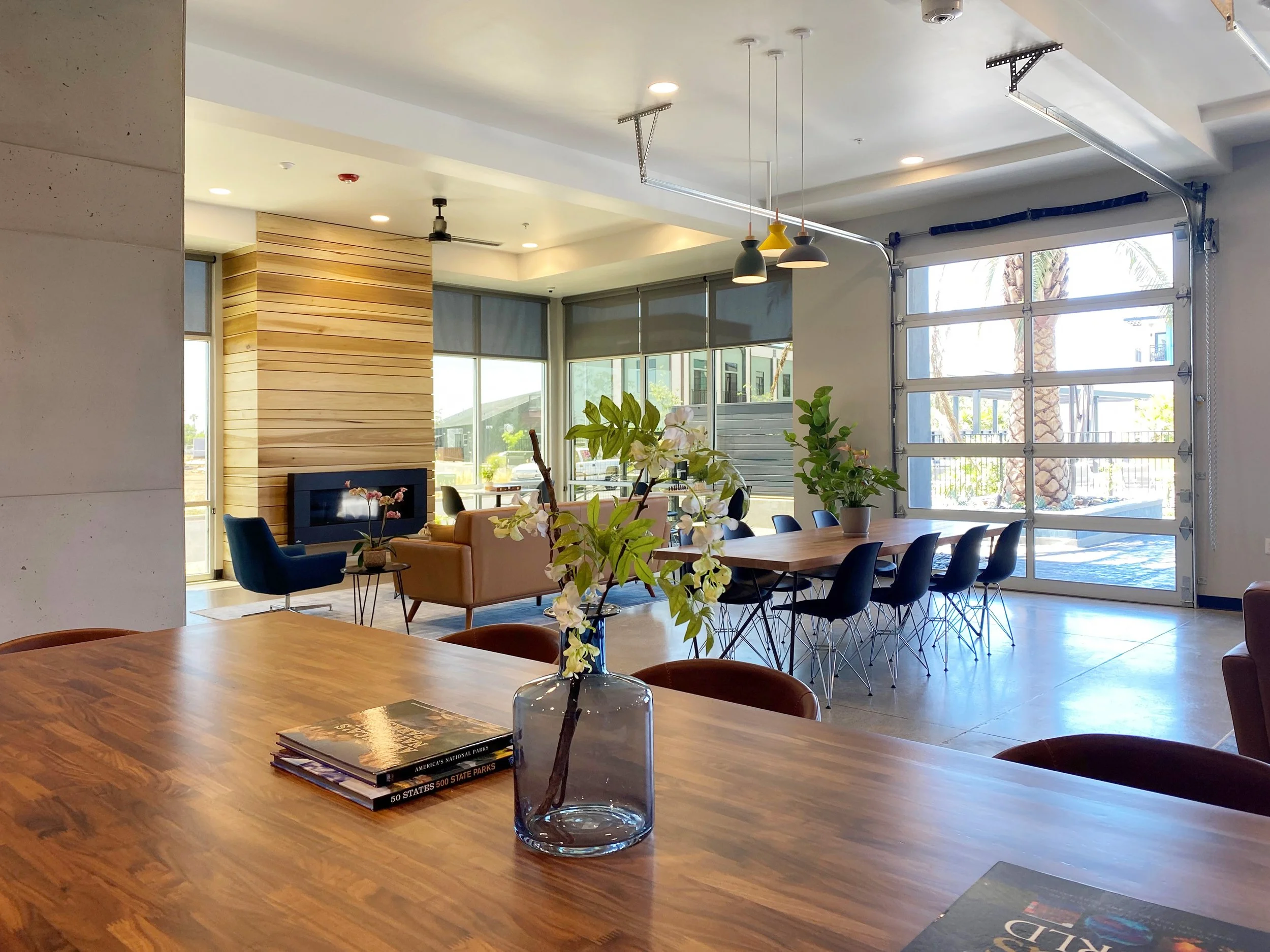 Modern indoor space with a large wooden table, vases with plants, and a spacious seating area featuring armchairs and a sofa near windows and glass garage-style door.
