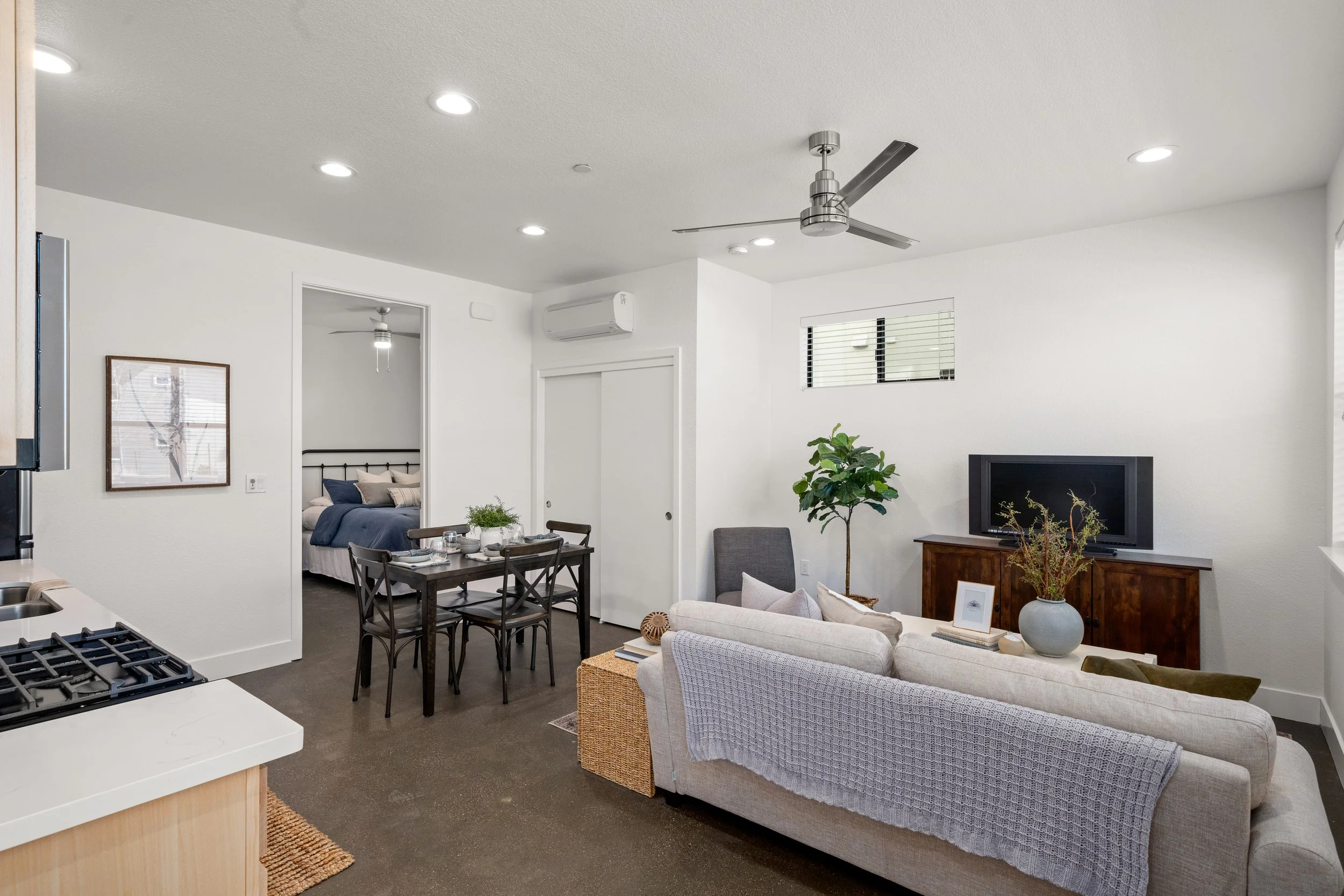 Modern living room with white walls, a beige sofa, a small dining table with black chairs, a television on a wooden stand, a gray armchair, a large potted plant, and a bedroom visible through an open doorway.